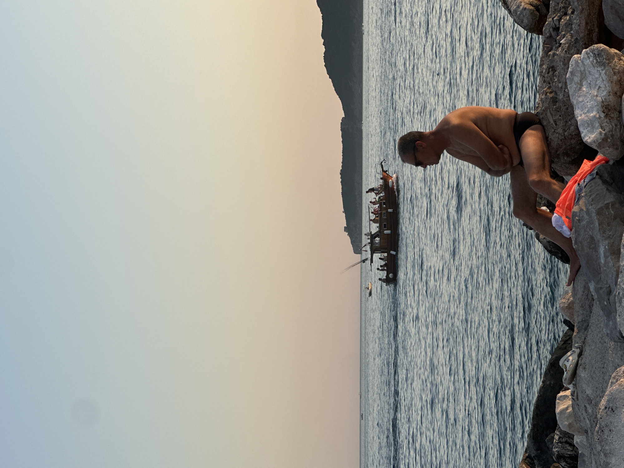 A man in black swim trunks sits on a rocky shore overlooking the Adriatic Sea in Rafailovići, Montenegro. The man is shirtless, wearing sunglasses, and has his arms crossed. In the middle ground, a wooden boat filled with passengers sails on the water. In the background, a dark, forested hill rises from the sea. The sky is a pale gradient of blue and orange, suggesting either dawn or dusk. The tide is low, exposing a line of weathered rocks at the water's edge where the man is seated.