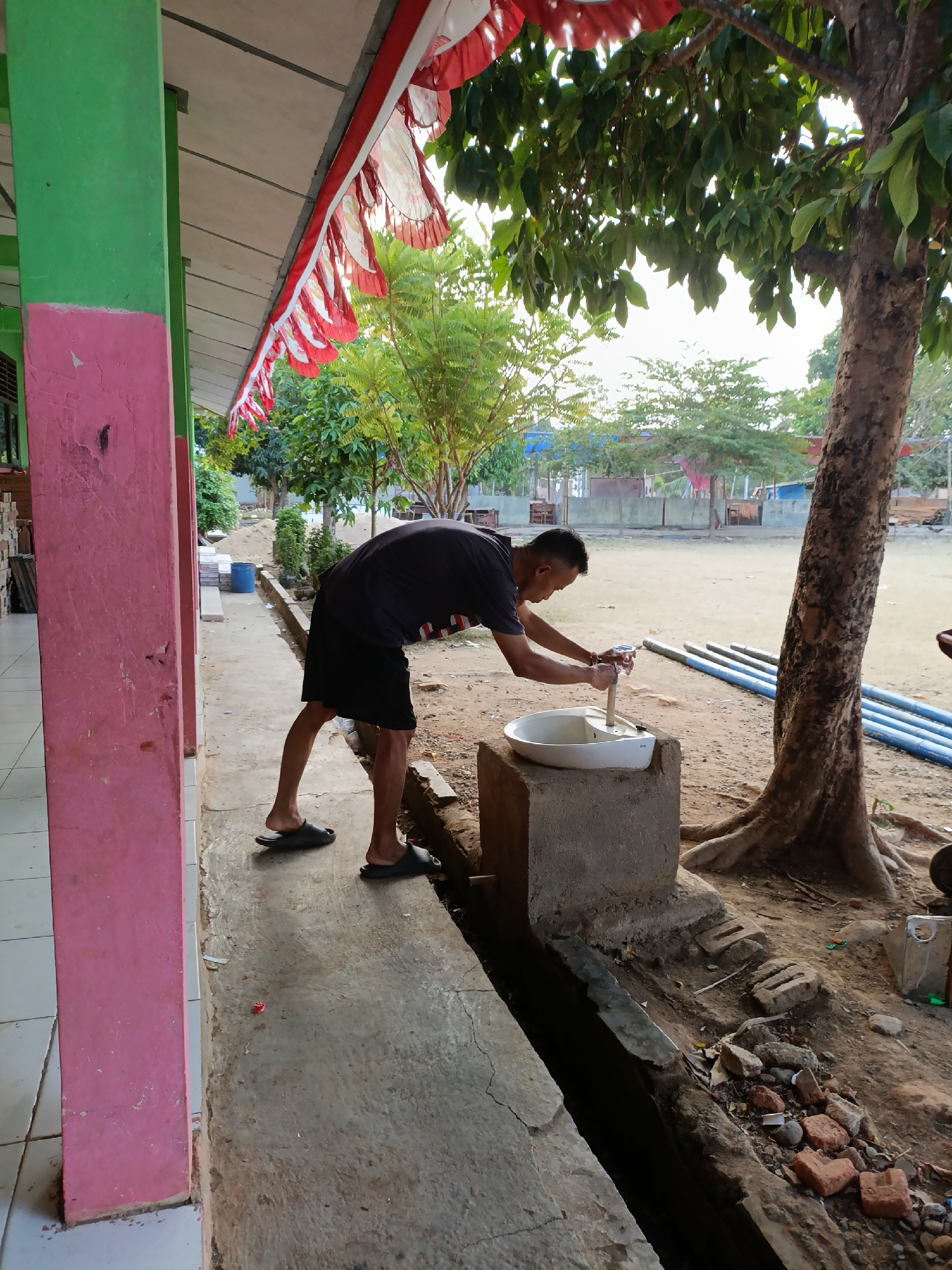 In the city of Sumberejo, Indonesia, a young man is seen at an outdoor sink. He is wearing a black t-shirt and shorts, and is leaning over the sink, with his hands on the faucet. The sink is a white ceramic basin mounted on a concrete pedestal. Behind the sink, a large tree trunk is visible. The scene appears to be in an outdoor area with a concrete walkway and a dirt ground. In the background, there are some buildings and trees. The overall impression is that of a simple, functional outdoor space.