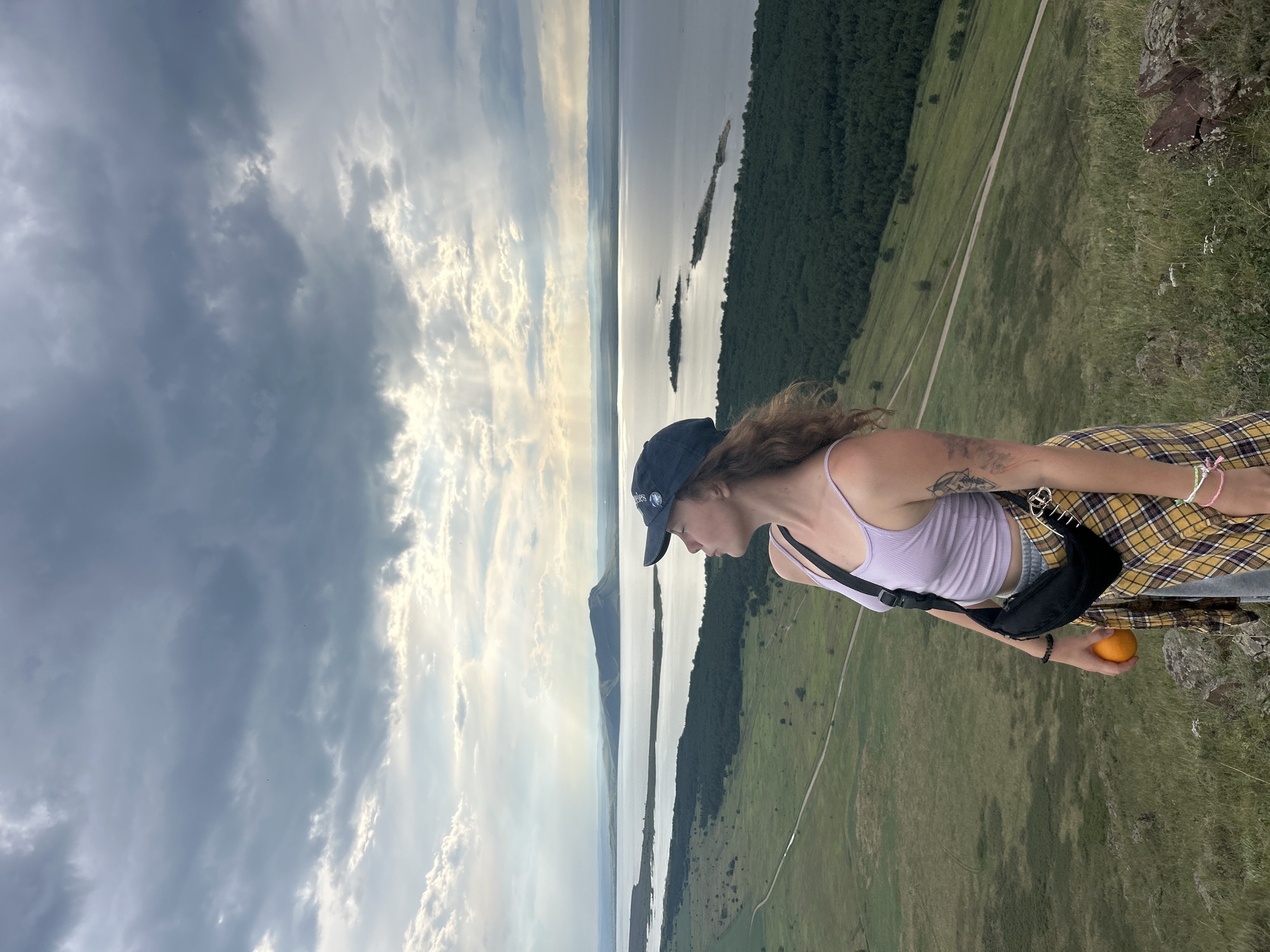 A young woman stands on a grassy hilltop, looking towards a vast body of water under a cloudy sky. She is wearing a light purple tank top, a yellow and black plaid shirt tied around her waist, and grey sweatpants. A black fanny pack is worn across her chest. She holds an orange in her right hand. Her left arm is bent at the elbow, and her left hand rests on her hip. A tattoo is visible on her upper left arm. She wears a dark blue baseball cap.
The background features a wide expanse of water, with a long, narrow island and several smaller ones scattered across it. A distant mountain range is visible on the horizon, and sunbeams break through the clouds, illuminating parts of the water and landscape. The foreground is a grassy slope dotted with rocks and sparse vegetation. A dirt road winds its way down the hill and across a green plain. The scene appears to be in Nikolo-Petrovka, Russia, a location known for its natural beauty and scenic views of water bodies and surrounding landscapes.