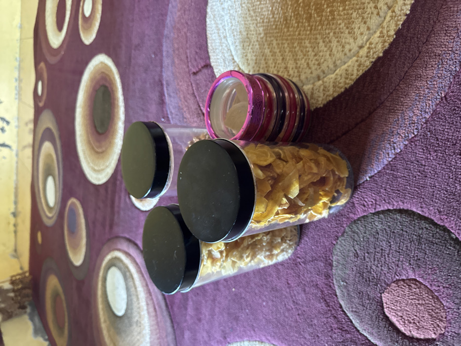 The image displays an indoor scene in the city of Pante, Indonesia. Three clear plastic cylindrical containers with black screw-top lids are arranged on a floor. The foremost container, positioned centrally, is filled with golden-yellow, irregularly shaped, flaky food items. Behind and to its left, a second container holds smaller, lighter beige, granular food items. A third container, partially visible in the upper right, also contains lighter granular items. Adjacent to the front container on the right, a stack of approximately 5-6 colored bangles, primarily in shades of pink, purple, and red, rests. The objects are situated on a purple-colored rug featuring large concentric circular patterns in various tones of purple, brown, beige, and white. To the right, a section of a light beige or cream-colored woven mat is visible. The background consists of a textured, light yellow wall. No individuals are present.