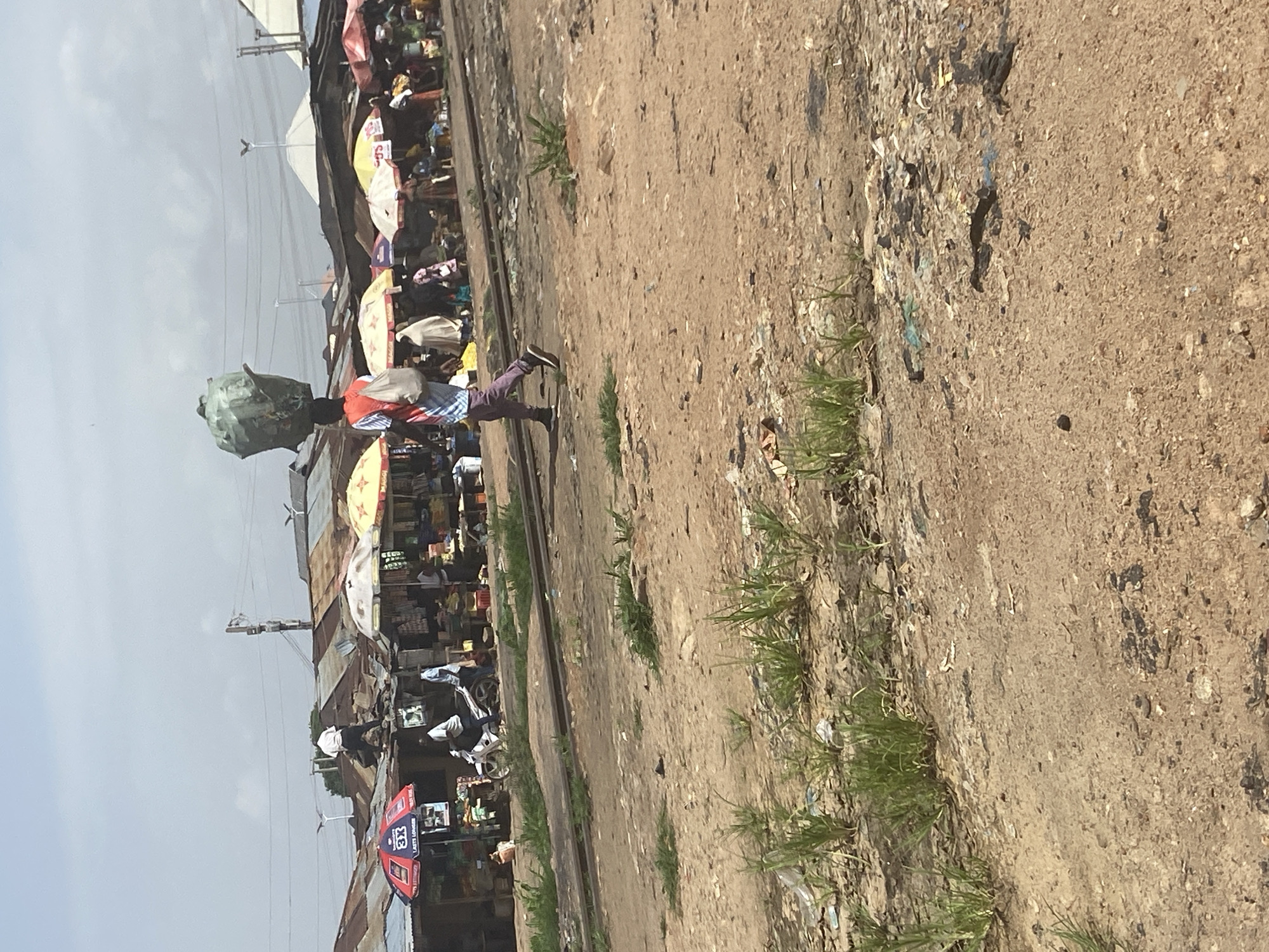 In Bukuru, Nigeria, a street market thrives under a hazy sky. A person walks across a dusty, uneven terrain, carrying a large green bundle balanced on their head and a bag slung over their shoulder. The market stalls, covered by colorful umbrellas, line a slightly raised area with rusty corrugated metal roofs. Some stalls display goods, while others appear to be under construction or in disrepair. A motorcycle is parked near one of the stalls, with two individuals standing nearby. In the foreground, dry soil and sparse vegetation are visible, with scattered debris. A set of railway tracks runs horizontally across the mid-ground, dividing the scene. The overall impression is of a bustling, utilitarian marketplace in a somewhat neglected urban setting.