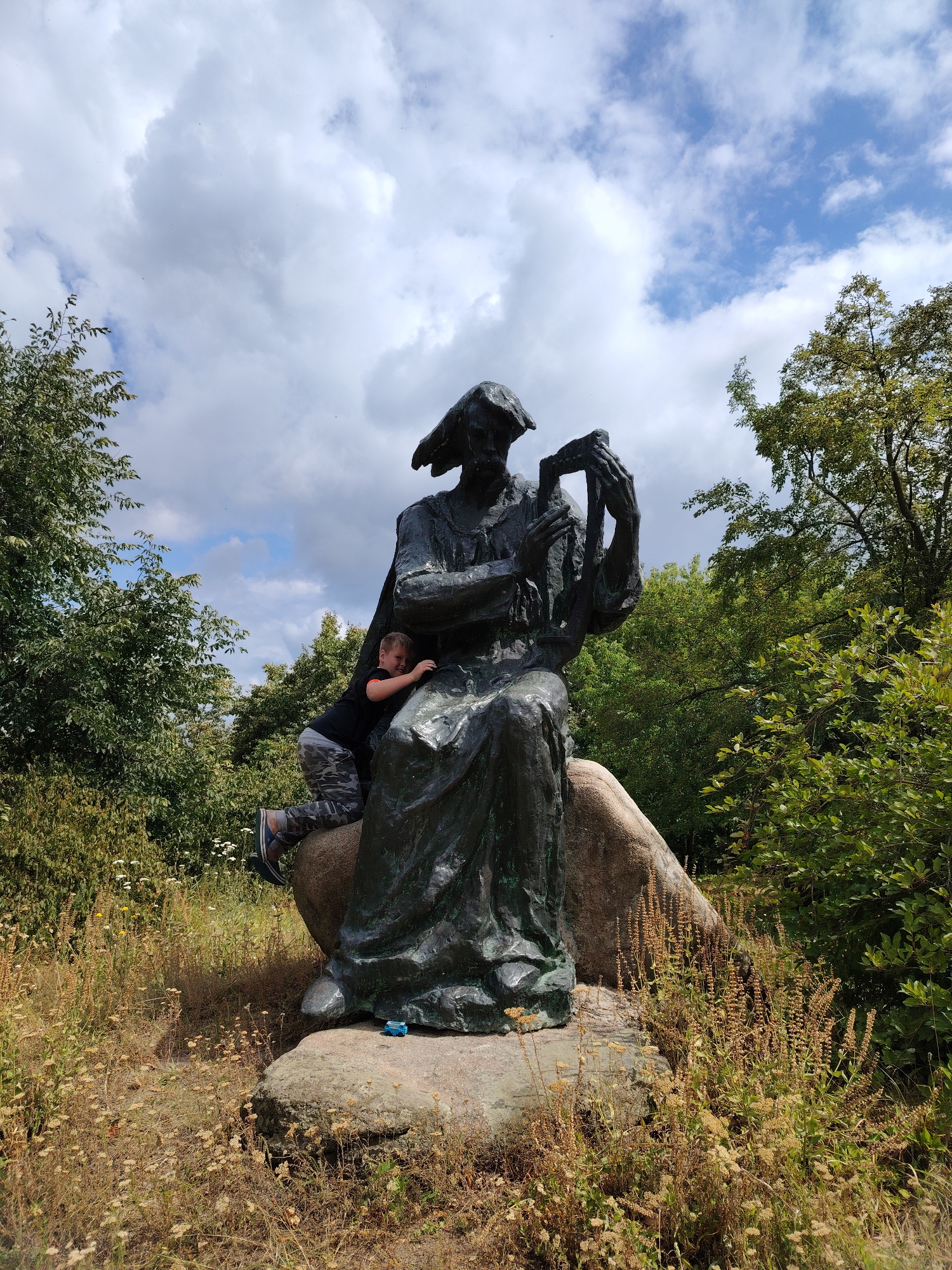 In Balyko-Shchuchynka, Ukraine, a young boy is pictured climbing on a large bronze statue situated outdoors amongst lush greenery and a boulder. The statue depicts a figure seated with a harp-like instrument. The boy, wearing camouflage pants and a black shirt, is positioned on the side of the statue, his body contorted as he grips the sculpture for support. The surrounding environment is wild, with dry grasses and wildflowers growing around the base of the statue and boulder. Trees and foliage frame the scene, with a cloudy sky overhead. A small blue toy is visible on the boulder at the base of the statue.