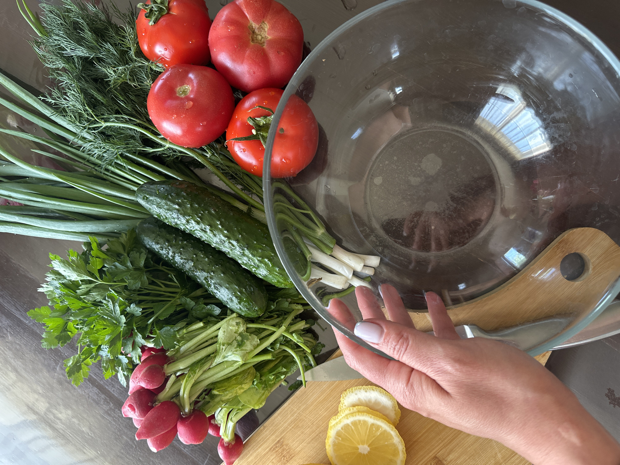 A still-life photograph captures a collection of fresh produce arranged around a clear glass salad bowl. The produce includes tomatoes, cucumbers, spring onions, dill, parsley, and radishes, all seemingly washed and ready for preparation. A wooden cutting board with lemon slices sits in the lower right corner, partially covered by a human hand. The scene appears to be set indoors on a dark-colored surface, possibly a kitchen countertop. The image does not contain information indicating that this image was taken in Chaikovsky, Russia.