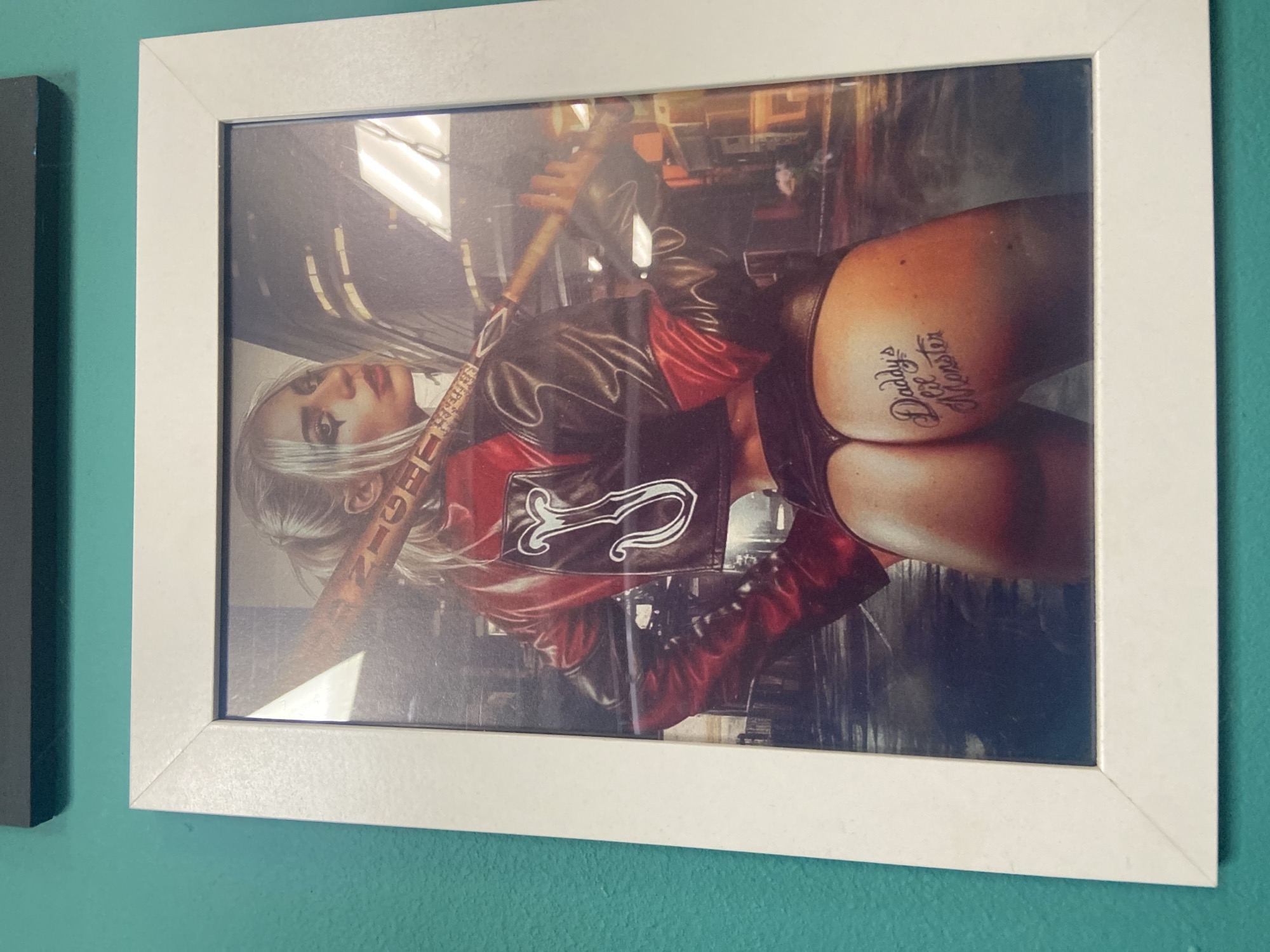 The image shows a framed print hanging on a teal-colored wall. The print depicts a stylized image of a woman in a red and black outfit, holding a baseball bat. The woman has blonde hair styled in a ponytail, and is wearing dark eye makeup and red lipstick. Her outfit includes a jacket with red sleeves and a black collar, and a dark thong. There is a tattoo on her buttock that reads "Daddy's lil Monster". The background of the print appears to be an urban setting at night, with buildings and streetlights. The print is framed in a white frame. The location in the city of Yegorlykskaya, Russia is not ascertainable from the image.