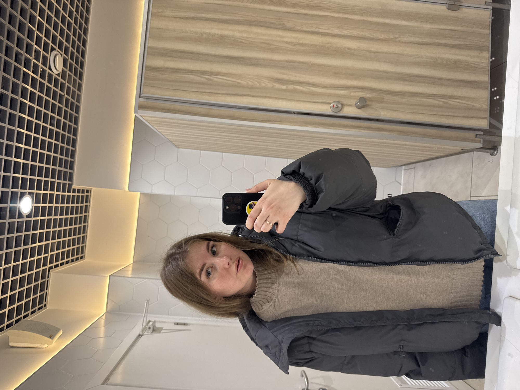 The photograph appears to be a selfie taken in a public restroom. The subject is a woman with light brown hair wearing a tan sweater and a black puffer jacket. She is holding a black smartphone in her right hand, presumably to take the photo.
The restroom features a combination of white hexagonal tiles on the walls and a wooden-look stall partition. The countertop and sink area are white, with a black grid-like pattern on an adjacent wall. There is a lighting fixture with a bright, white light that illuminates the scene.
There is no indication of the location being Perm, Russia, or any identifying markers within the photograph. Therefore, I cannot confirm or deny that the location is in Perm, Russia.