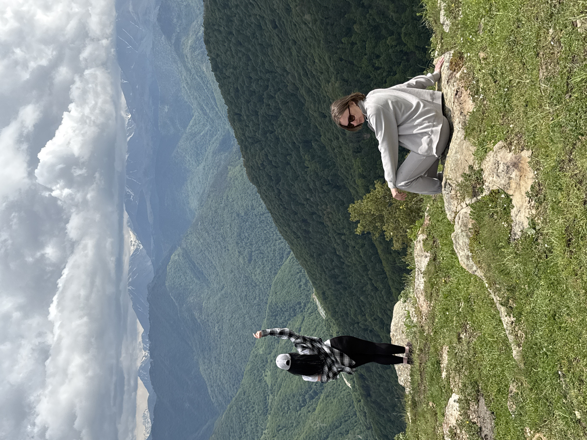 Two individuals are situated on a rocky, grassy overlook with a mountainous backdrop. On the left, a person stands with their back to the camera, wearing a white baseball cap, a white t-shirt, a black and white plaid shirt tied around their waist, and black leggings. Their right arm is raised, pointing upwards. On the right, a second person is seated on a rock, facing left, wearing a light gray tracksuit and sunglasses. They are looking away from the camera. The landscape behind them consists of numerous green, tree-covered mountain ranges that recede into the distance, with snow-capped peaks visible on the horizon under a cloudy sky. The scene appears to be in a natural, elevated location, possibly a viewpoint or hiking spot, consistent with the mountainous terrain of Нижняя Теберда, Russia.