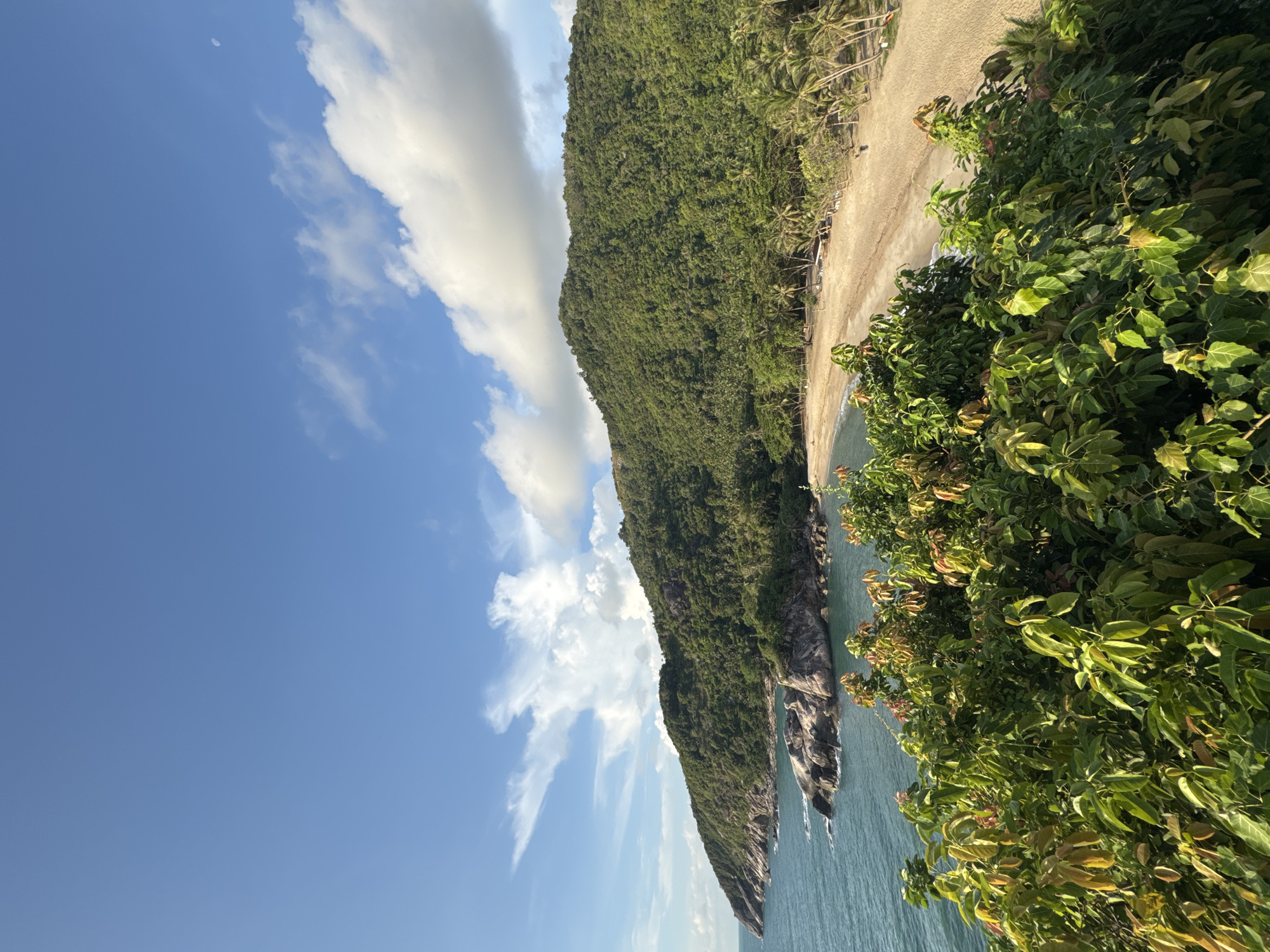 Captured from an elevated position, the image showcases a coastline scene near Baan Thong Nang, Thailand. A forested hill dominates the landscape, running parallel to a sandy beach. The beach is lined with sparse vegetation and a few structures. The ocean meets the shoreline, with some rocky outcrops visible. Lush green foliage fills the foreground. The sky is blue with scattered clouds. A small moon is visible in the sky.