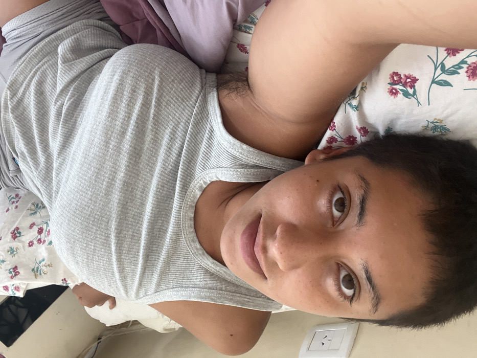 An adult individual is lying supine on a bed, captured in a close-up, top-down perspective. The subject has short dark hair, light brown skin, and is looking directly at the camera with wide brown eyes. They are wearing a light grey, ribbed tank top and partially visible grey shorts. The left armpit shows visible hair. The bed includes a white pillow with a floral pattern (pink/purple flowers, green leaves), a plain white pillow, and purple bedding. On the light-colored wall behind the subject's head, a white Type I electrical outlet with three angled flat pins is visible. The scene depicts a domestic interior, consistent with a private residence in Buenos Aires, Argentina.