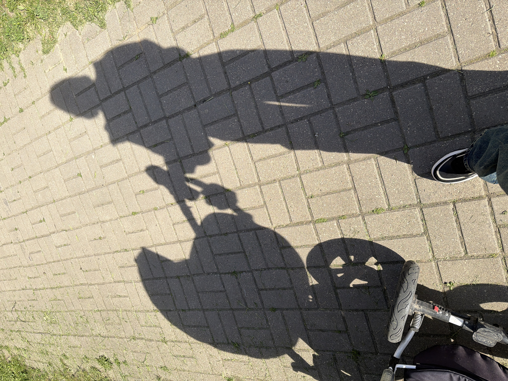 The image shows the shadow of a person pushing a stroller on a brick walkway in an unknown city. The walkway is made of light grey bricks. A small amount of grass is visible on the edge of the walkway. A person wearing jeans and black sneakers is standing near the stroller. The shadow cast by the stroller indicates a wheel, and the shadow of the person pushing it is also visible. The lighting suggests it is daytime.
