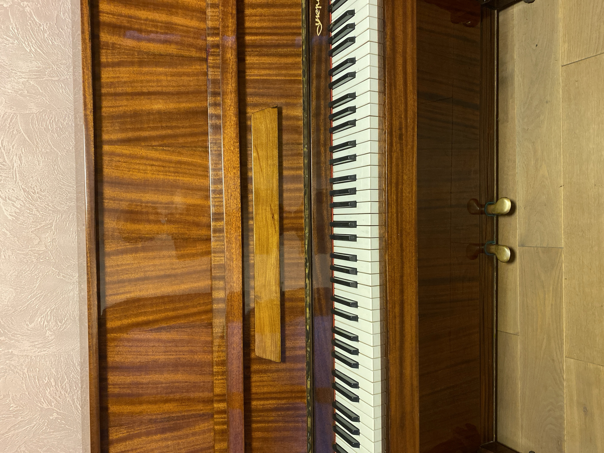 The image shows a close-up, eye-level view of the lower half of an upright piano. The piano is made of polished wood with a rich, reddish-brown grain. The keyboard features white and black keys, with the black keys appearing to be slightly worn. Above the keyboard, a wooden plank is affixed, and above that, the polished wood of the piano continues upwards. The top edge of the piano is visible, meeting a textured, light-colored wall. Below the keyboard, two brass pedals are visible, resting on a wooden floor. The floorboards are light brown and laid horizontally. The overall scene suggests an indoor setting, likely a room in a home. The lighting is even, highlighting the sheen of the wood. There are no people or animals in the image.