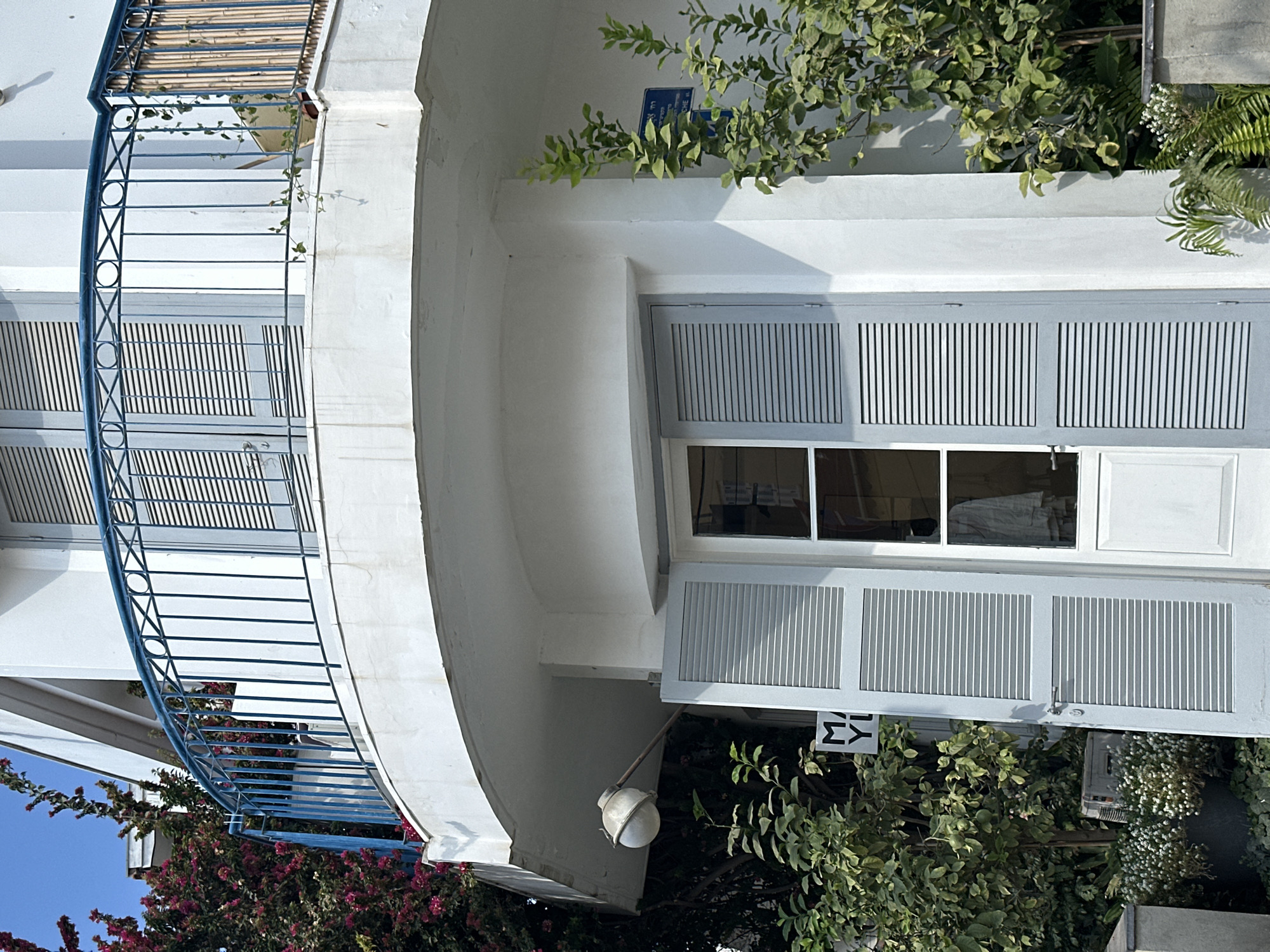 The image depicts the exterior of a building, likely a residential or commercial property, in Tel-Aviv, Israel. The architecture features a curved balcony with a blue metal railing adorned with circular and criss-cross patterns. Below the balcony, a white wall curves outwards, and a white door with light grey shutters is visible. The shutters are partially open, revealing a window behind them. Green foliage, including bushes and trees, surrounds the lower part of the building. A sign with the letters "MYL" is affixed to a pole near the door. In the background, a vibrant pink flowering plant cascades down the side of the building, and a blue sky is visible. The scene suggests a sunny day in an urban environment.