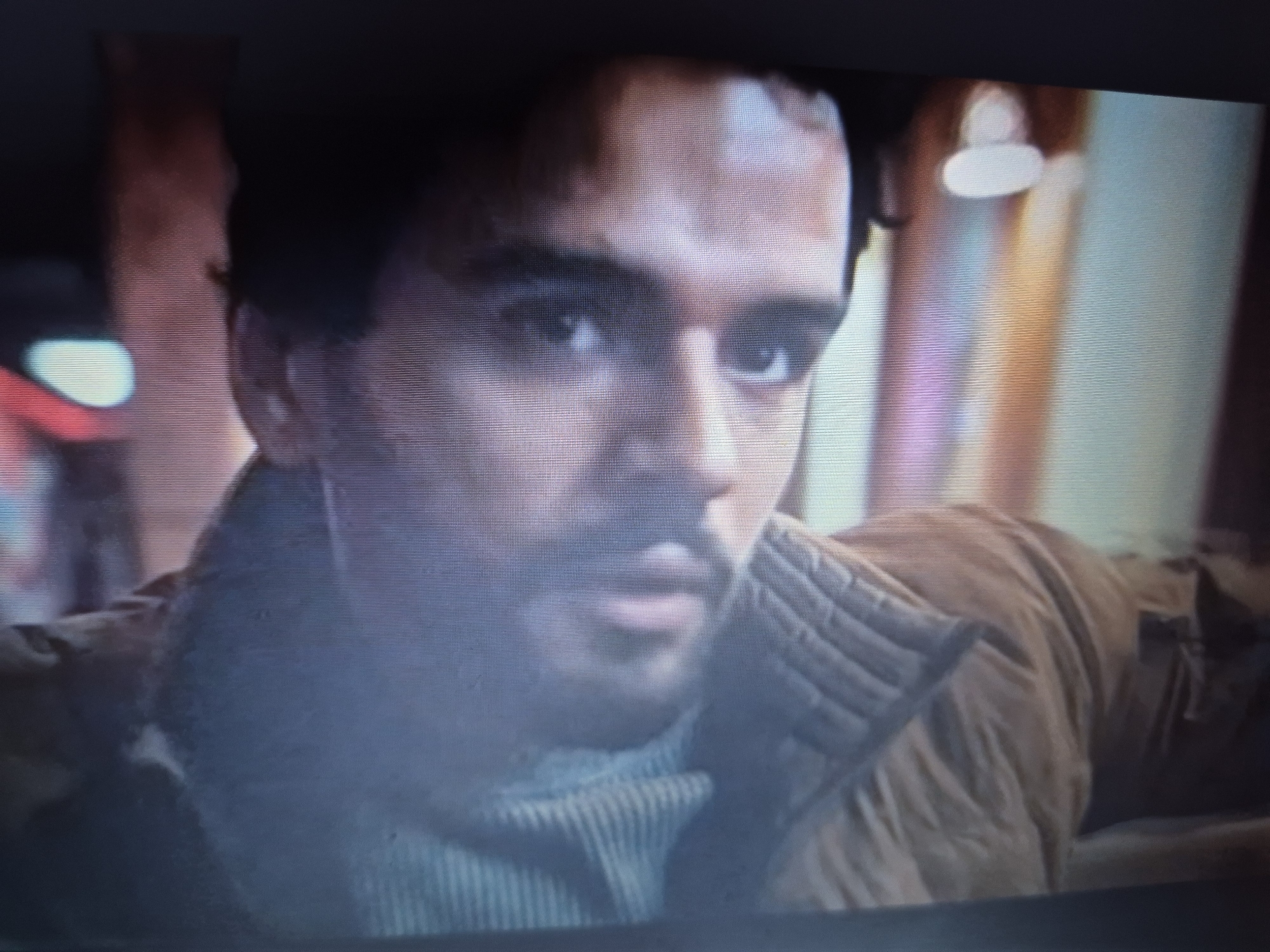 The image is a close-up, low-resolution photograph of a man's face. The man has dark hair and is looking directly at the camera with a neutral expression. He is wearing a light-colored, ribbed turtleneck sweater and a brown, quilted jacket. The lighting is dim and appears to be artificial, with some blurry lights in the background that suggest an indoor setting. The image quality is poor, with visible scan lines and pixelation, giving it a grainy appearance. There is no discernible location in Sandwell, United Kingdom visible in the image.