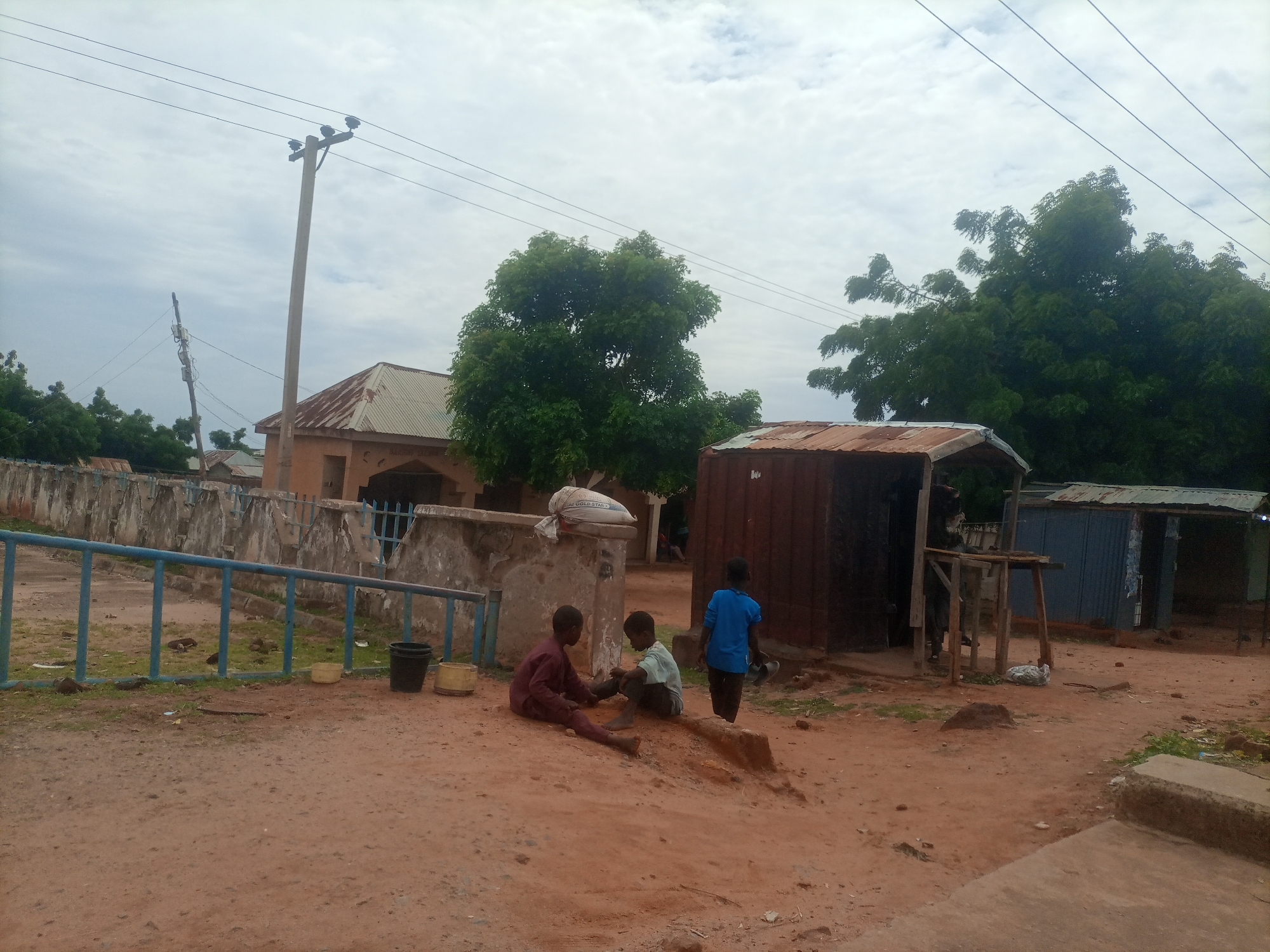 In the city of Wuro Dole, Nigeria, a dirt road leads past a building with a corrugated metal roof, a concrete wall, and a blue metal railing. Three children are present: two are sitting on the ground, one in a maroon garment, the other in a light green shirt and dark pants. The third child, in a bright blue shirt and dark pants, stands and faces away from the camera. Power lines crisscross the cloudy sky. Behind the children are several structures, including a weathered wooden stall and a building with a faded sign. The ground is covered in reddish-brown dirt and sparse vegetation.