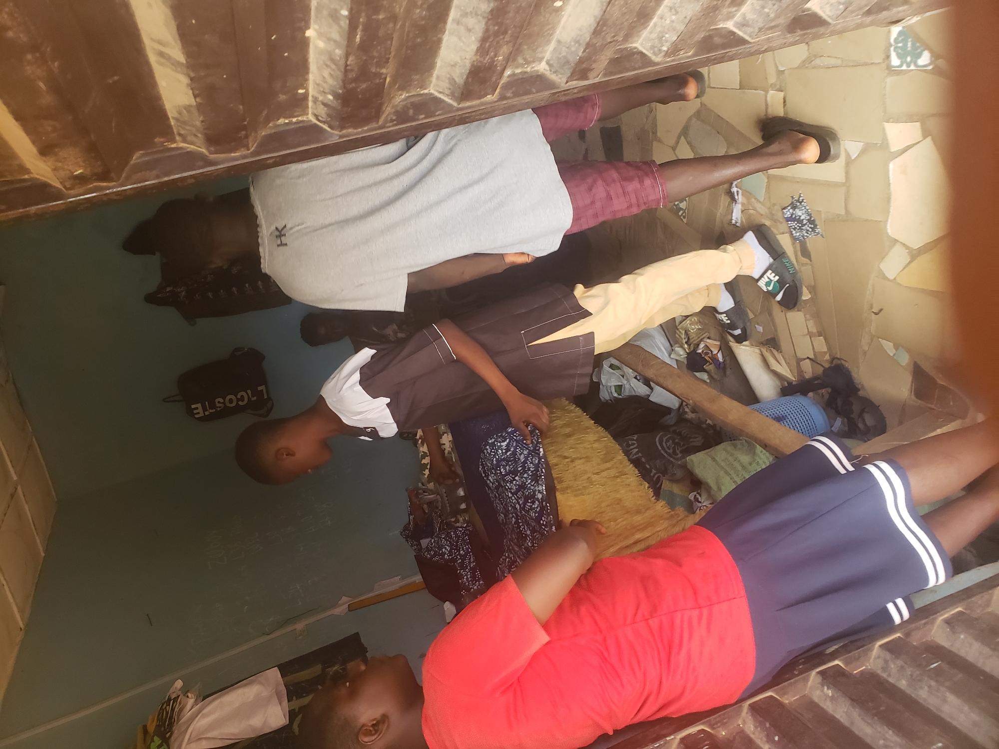In Kachia, Nigeria, a group of young people are gathered in a dimly lit room with blue walls. A young female wearing a red t-shirt and a navy blue pleated skirt stands facing a table laden with patterned fabrics. To her right, a young male in a brown and white tunic and cream-colored pants is interacting with the fabrics on the table. Further to the right, another young male in a grey t-shirt with an "HK" logo and maroon checkered shorts stands partially obscured by a wooden doorway. The room appears to be a workshop or tailor's shop, with clothes hanging on the wall and a cluttered floor with scattered tiles and debris.