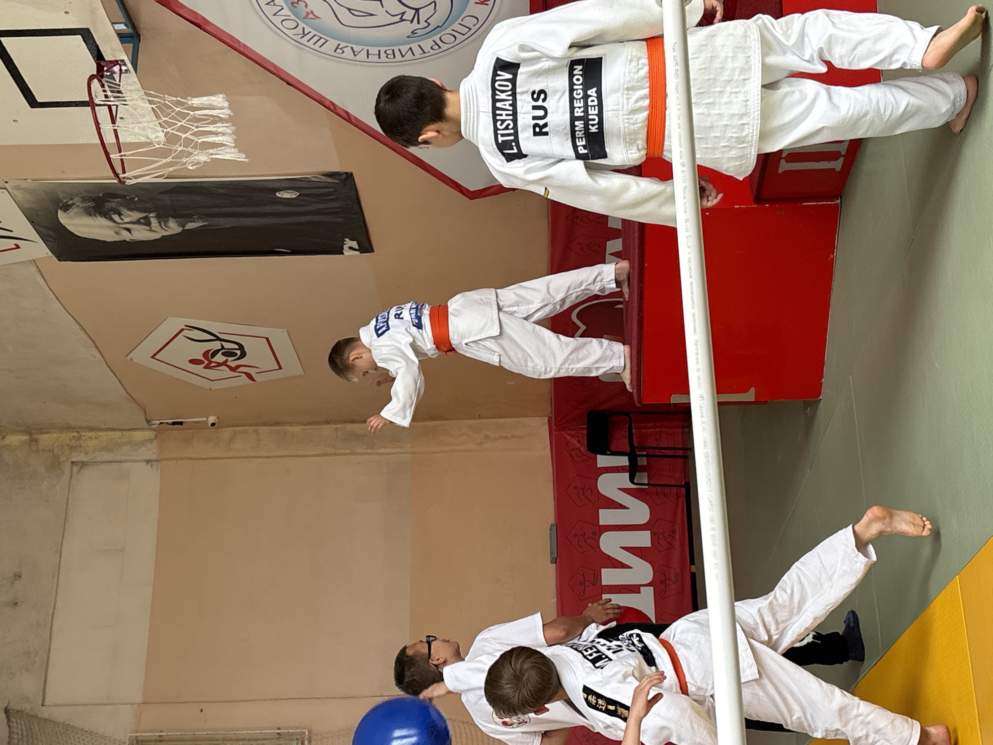 The image captures an indoor martial arts training session in a gymnasium located in Kuyeda, Perm Region, Russia. Three young boys, all wearing white judogis with orange belts, are visible. The boy in the center-right, facing away from the camera, has "L.TISHAKOV RUS PERM REGION KUEDA" emblazoned on his back. He stands on a green mat. Another boy, positioned on a red platform to the left, is in a ready stance with raised hands. A third boy is partially visible in the foreground-left, moving across a yellow mat. An adult male, wearing a white t-shirt and glasses, stands on the green mat near the red platforms, appearing to supervise. The background features two-toned walls (light beige and peach/brown). Hanging on the walls are a large black and white portrait of an elderly Asian man, a white hexagonal emblem with a red stylized figure, and a partially visible banner with the Cyrillic text "СПОРТИВНАЯ ШКОЛА" (Sports School). A basketball hoop with a white net is mounted on the right wall, above a red and white banner. A white dome security camera is affixed high on the right wall. A blue ball is partially visible near the adult.