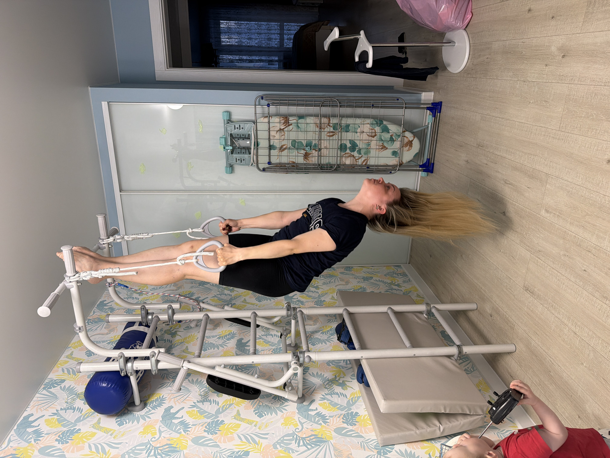 A woman is performing a gymnastic exercise on a white metal structure with a padded base. She is suspended horizontally, holding onto white rings with ropes attached above her. Her body is extended, with her feet near the top of the structure and her head towards the other end. She is wearing a dark t-shirt and dark shorts. Her blonde hair is flowing downward.
Behind her is a wardrobe with sliding doors, above which is an ironing board on a rack. In the background, a clothing rack and a plastic bag can be seen. The room has light-colored wooden flooring and wallpaper with a floral pattern.
In the foreground, at the bottom of the image, a child in a red shirt is lying on the padded base of the structure. The child is holding a small black device.
There is no information to confirm the location of the image in Votkinsk, Russia.