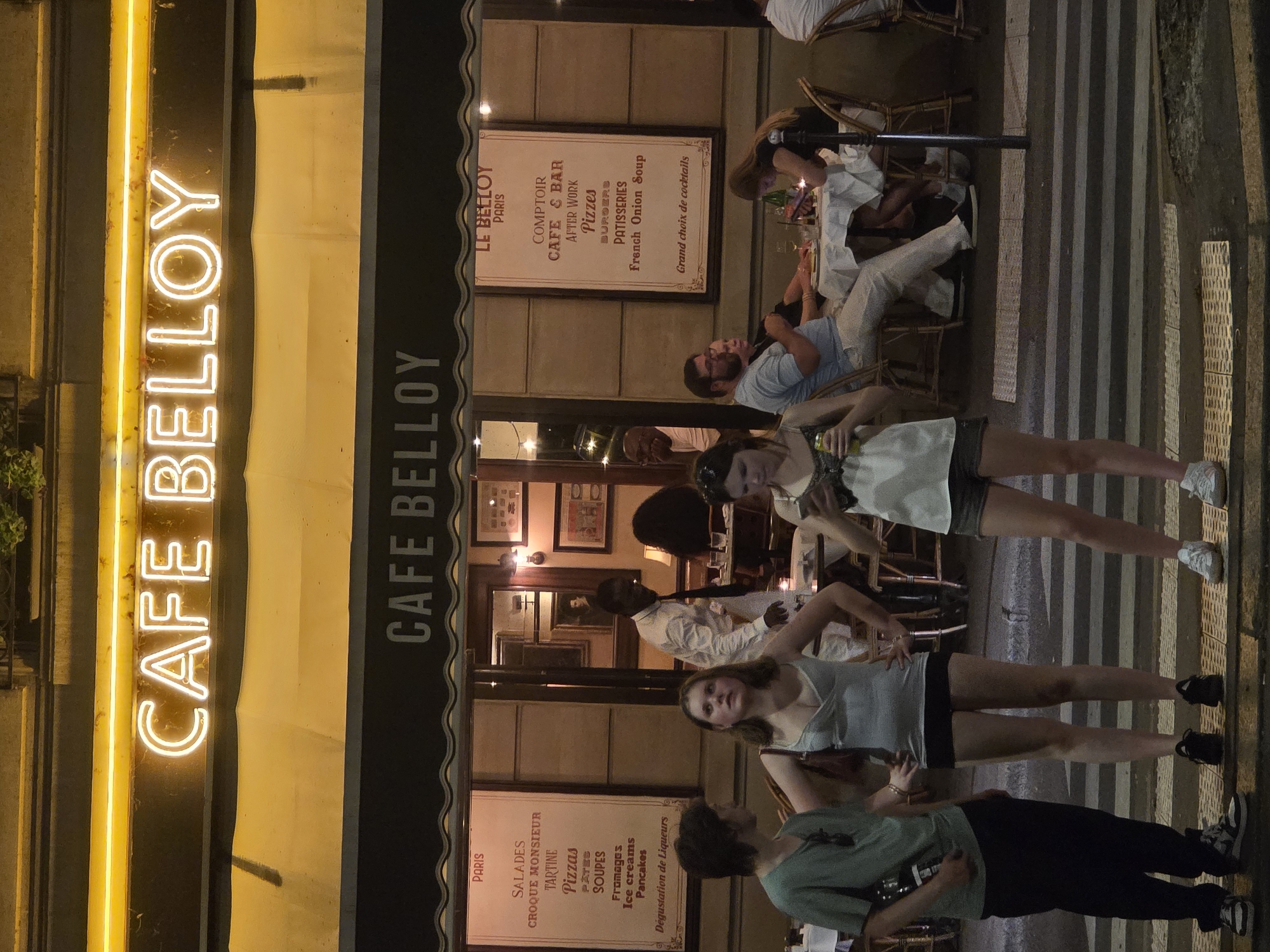 This is a nighttime, eye-level, medium shot of the exterior of a cafe in Paris, France. The cafe is named "CAFE BELLOY" and is illuminated by a large neon sign above the entrance and smaller signs displayed inside. The facade of the cafe is light brown with large windows. Several tables with chairs are set up outside the cafe. People are seen standing and sitting at these tables, some of whom appear to be patrons. The cafe is situated on a street with a pedestrian crossing in the foreground. The lighting is dim, with the neon signs providing the primary illumination.