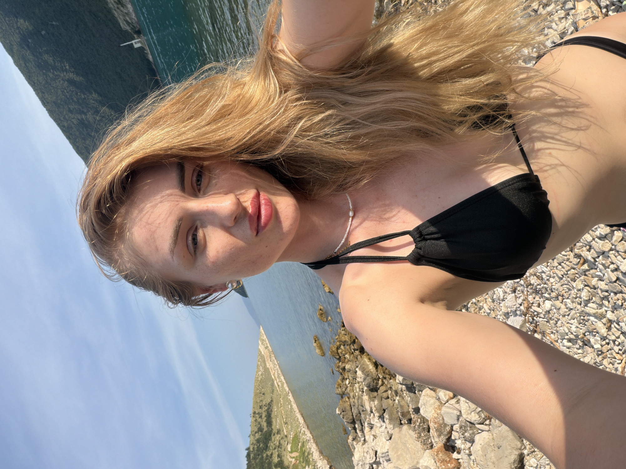 A young woman with long blonde hair is lying on a rocky beach, taking a selfie. She is wearing a black bikini and a pearl necklace. The sun is shining, casting shadows on her skin. Behind her, the turquoise water of the sea stretches out to the horizon, with a green, tree-covered hillside in the distance. The sky is a clear, bright blue. The overall impression is one of a relaxing summer day by the sea.