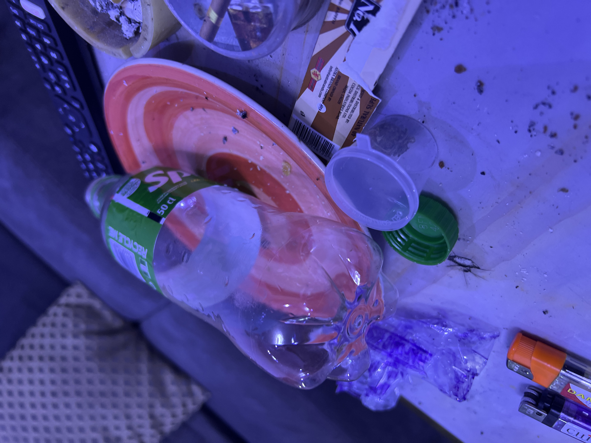 The image presents a messy scene with various objects scattered on a surface. A half-empty plastic bottle labeled "Sprite" (50 cl) with the "RECYCLE ME" message prominent lies partially on a plate with red and white concentric circles. A pack of rolling papers labeled "No. 1" is visible, indicating it is distributed by ICG Distributions LTD. in Lagos, Nigeria, with the brand owned by Astra Brands Company. Adjacent to the bottle are a clear plastic funnel, a green bottle cap, a small transparent bag with purple residue, and two lighters – one orange and one purple. The environment is cluttered and appears to be indoors. It is not possible to tell where it is from Abuja, Nigeria.