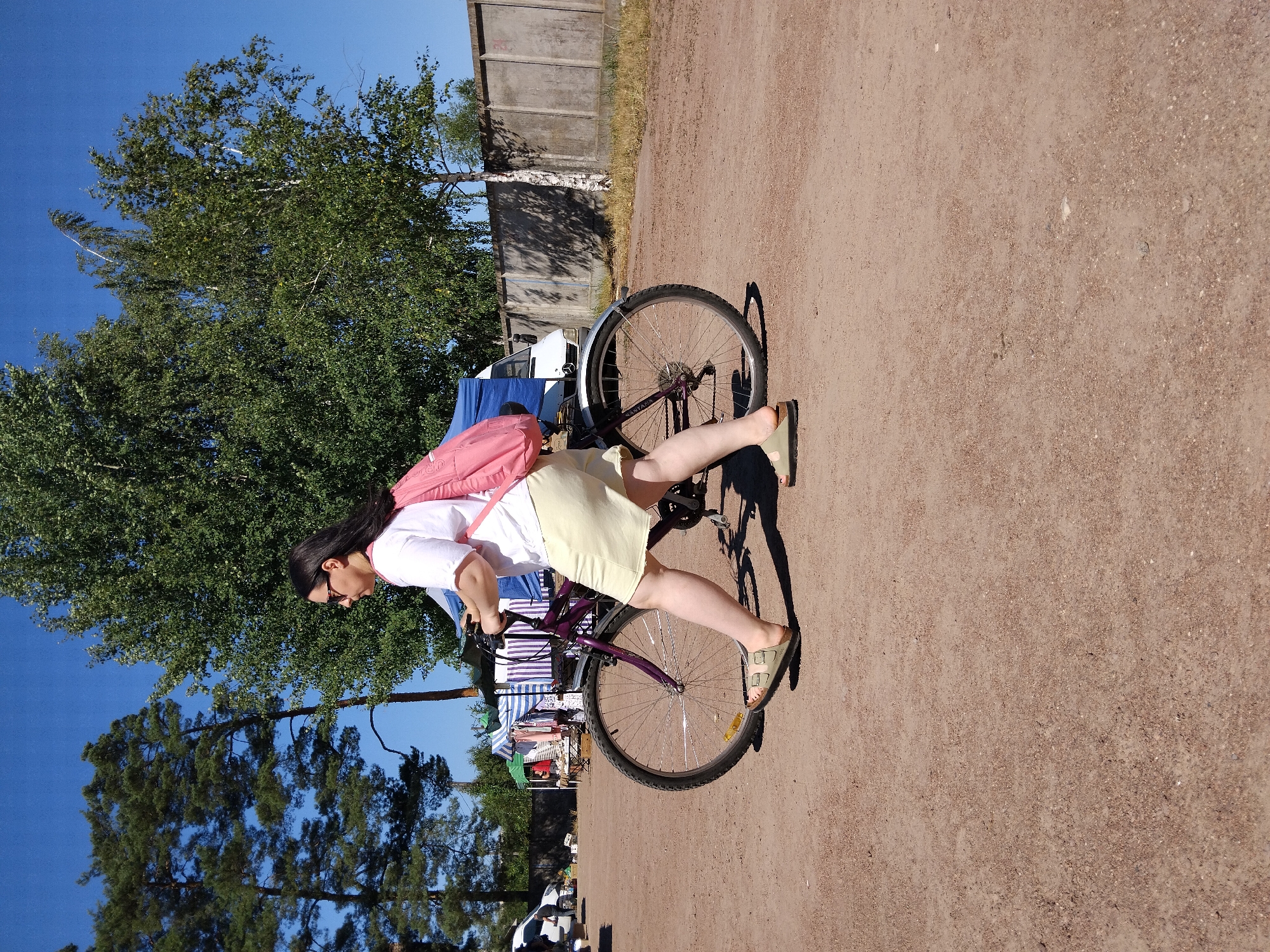 The image depicts a woman walking a bicycle on a dirt ground with a few trees and stalls in the background. She is wearing a white t-shirt, a light yellow skirt, and sandals, with a pink backpack on her shoulders. The bicycle is purple with a black seat and wheels. In the background, there are several stalls with striped awnings, suggesting a market or fair. The sky is clear and blue. The scene appears to be an outdoor event in the city of Irshansk, Ukraine.