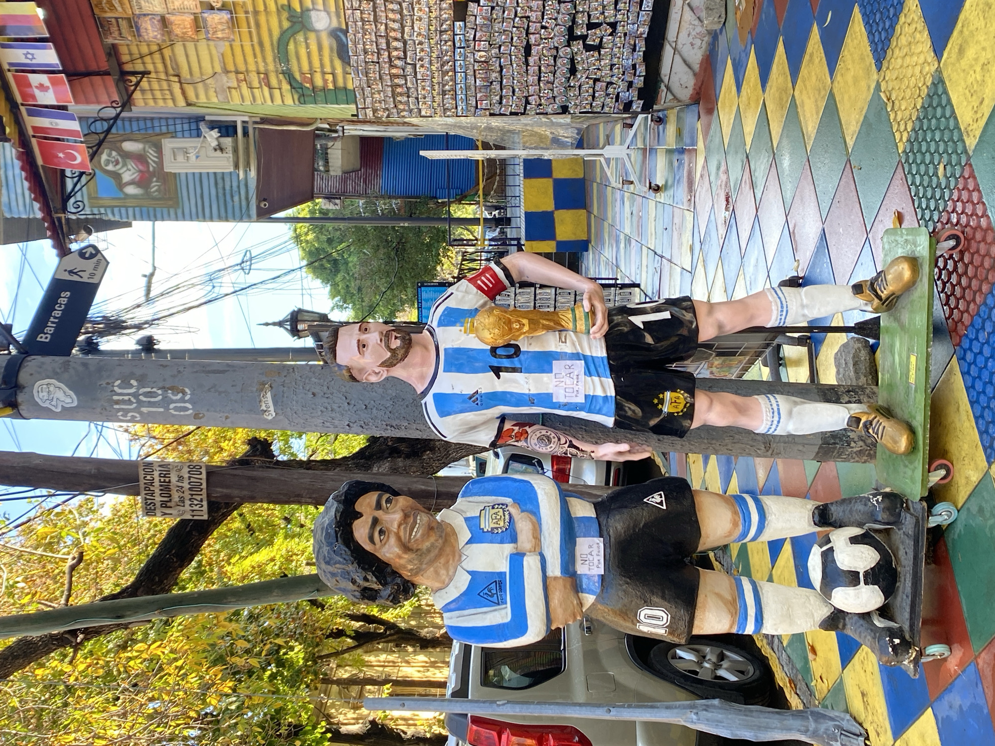 The image captures a vibrant street scene in Buenos Aires, Argentina, featuring two life-sized statues of iconic Argentine football players. On the left, a statue of Diego Maradona stands with crossed arms and a smiling expression, wearing a blue and white striped Argentina jersey with "10" on the back and the "AFA" and "Le Coq Sportif" logos. A soccer ball rests at his feet. To the right, a statue of Lionel Messi is depicted holding a replica of the FIFA World Cup trophy, wearing a light blue and white striped Argentina jersey with "10," "AFA," and "adidas" logos. Both statues have small signs that read "NO TOCAR POR FAVOR" (Do not touch, please).
The setting is an extremely colorful street, characteristic of areas like Caminito in La Boca. Buildings in the background are painted in bright, geometric patterns including yellow and blue checkerboards, diamond shapes, and solid colors. One building facade is adorned with numerous small, framed pictures or tiles. The ground beneath the statues is also painted in a multi-colored diamond pattern.
Notable details include a street sign on a utility pole pointing to "Barracas" with an arrow and "10 min," indicating a nearby location. Several national flags, including those of Israel, Canada, and Turkey, hang from a building. Other visible text includes "DESTAPACION Y PLOMERIA Las 24 hs 1132100708" (plumbing services) on another sign attached to a pole. The scene is brightly lit by daylight, suggesting good weather. The overall atmosphere is lively and celebratory, reflecting Argentina's passion for football.
