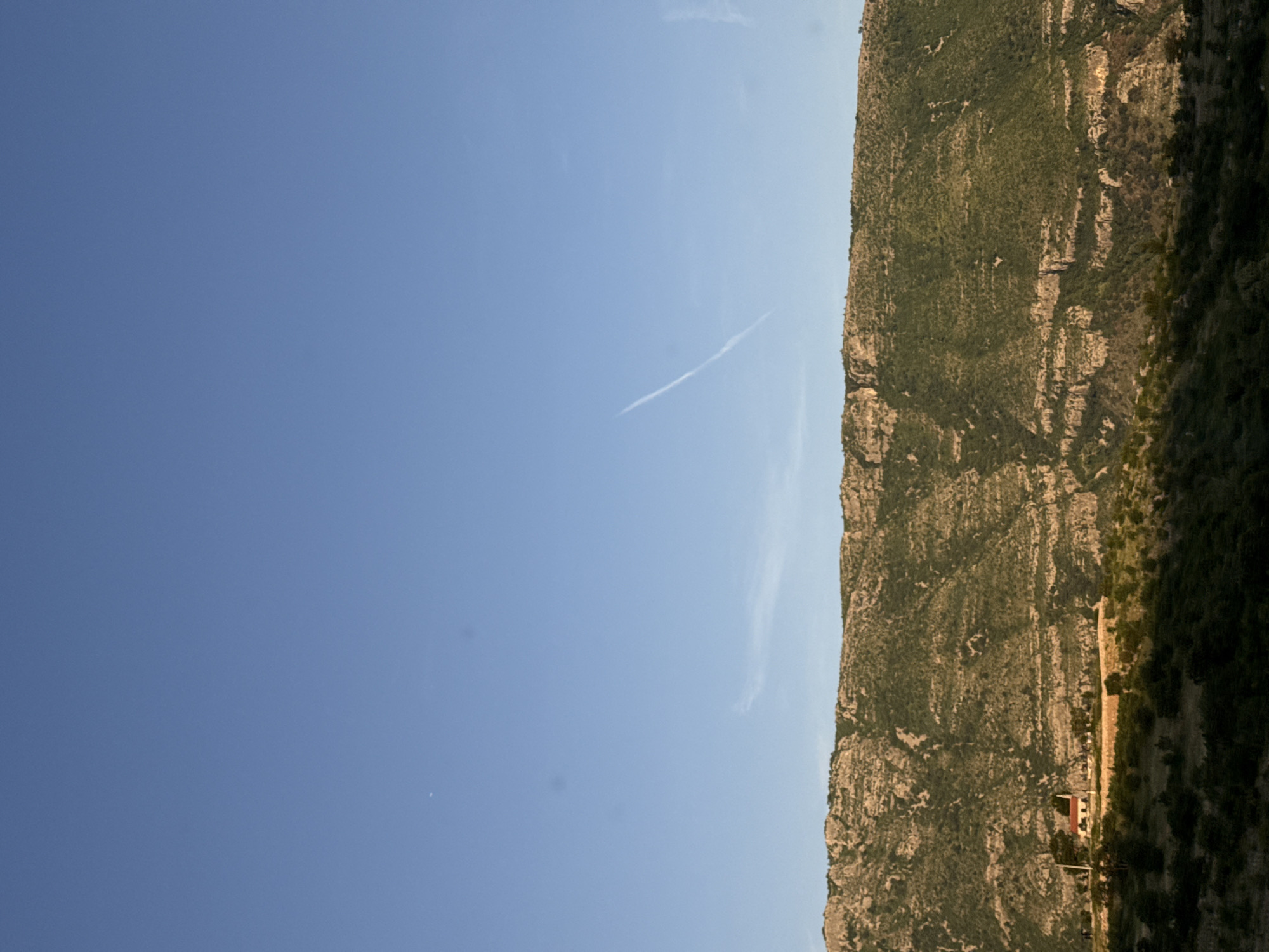 The image depicts a landscape, likely located in the vicinity of Klopot, Montenegro, featuring a clear blue sky on the left side and a densely vegetated, rocky mountainside on the right. There is a contrail visible in the upper center of the sky. Lower down the mountainside, a building with a red roof is discernible.