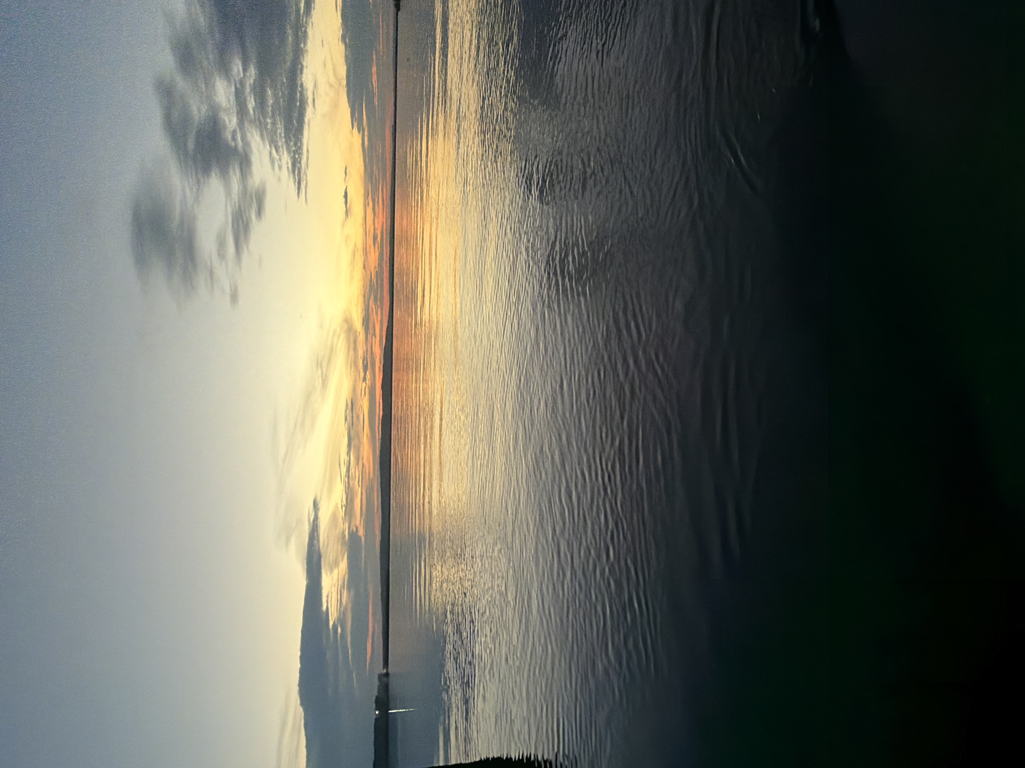 The image depicts a serene and atmospheric scene of a body of water, likely a lake or ocean, at sunset. The orientation of the photograph is vertical, with the water occupying the majority of the frame.
The sky is a dramatic blend of colors. On the left side, there are soft, diffused clouds, tinged with a pale grey. As the view moves towards the right, the sky explodes with the vibrant hues of sunset. Oranges, yellows, and hints of pink streak across the horizon, illuminating the wispy clouds and casting a warm glow.
The water surface reflects these sky colors, creating a shimmering, textured expanse. The right side of the image shows the water in deeper, darker tones, with subtle ripples creating a pattern of light and shadow. A thin, dark line runs horizontally across the frame, suggesting a distant landmass or shoreline. Near the bottom of the frame, there are hints of man-made structures, possibly lights from a distant town or boats, but they are indistinct.
The overall mood of the image is peaceful and contemplative, evoking the quiet beauty of twilight. The time of day is clearly sunset, with the low angle of the sun creating the characteristic warm lighting and dramatic sky. There are no discernible people or animals in the image. No text is visible.