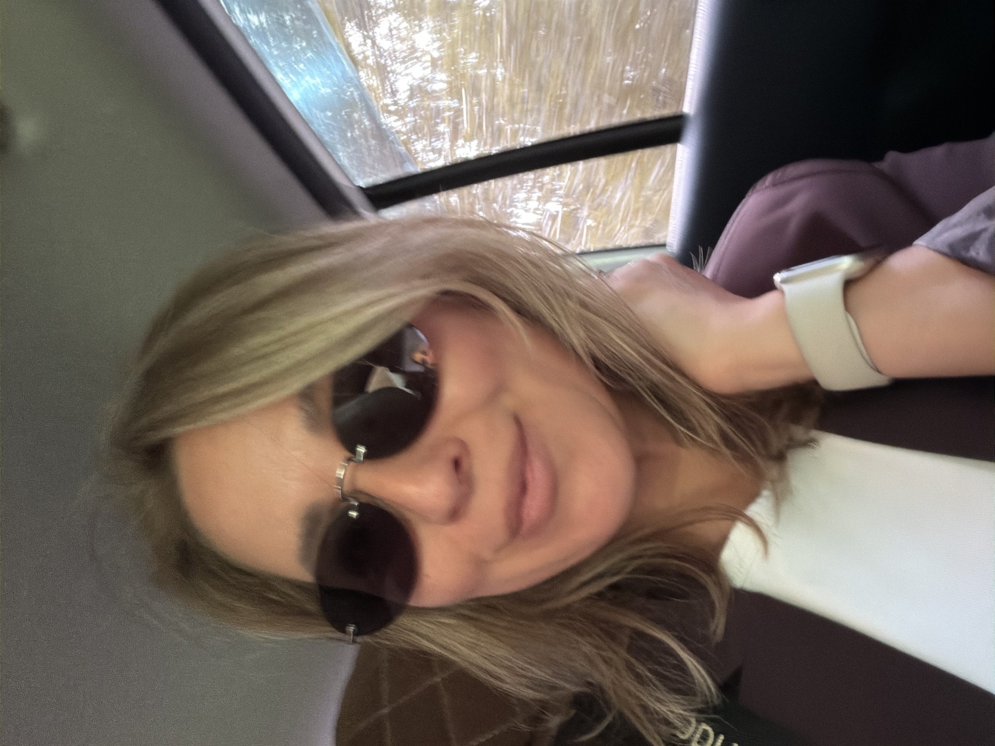 A woman with sunglasses and a smartwatch is sitting inside a vehicle, possibly a car, based on the window and seat visible in the background. The outside environment seems to be an area with trees, potentially a forest or park. The image was taken inside the vehicle. There is no information about the city of Chaikovsky, Russia.
