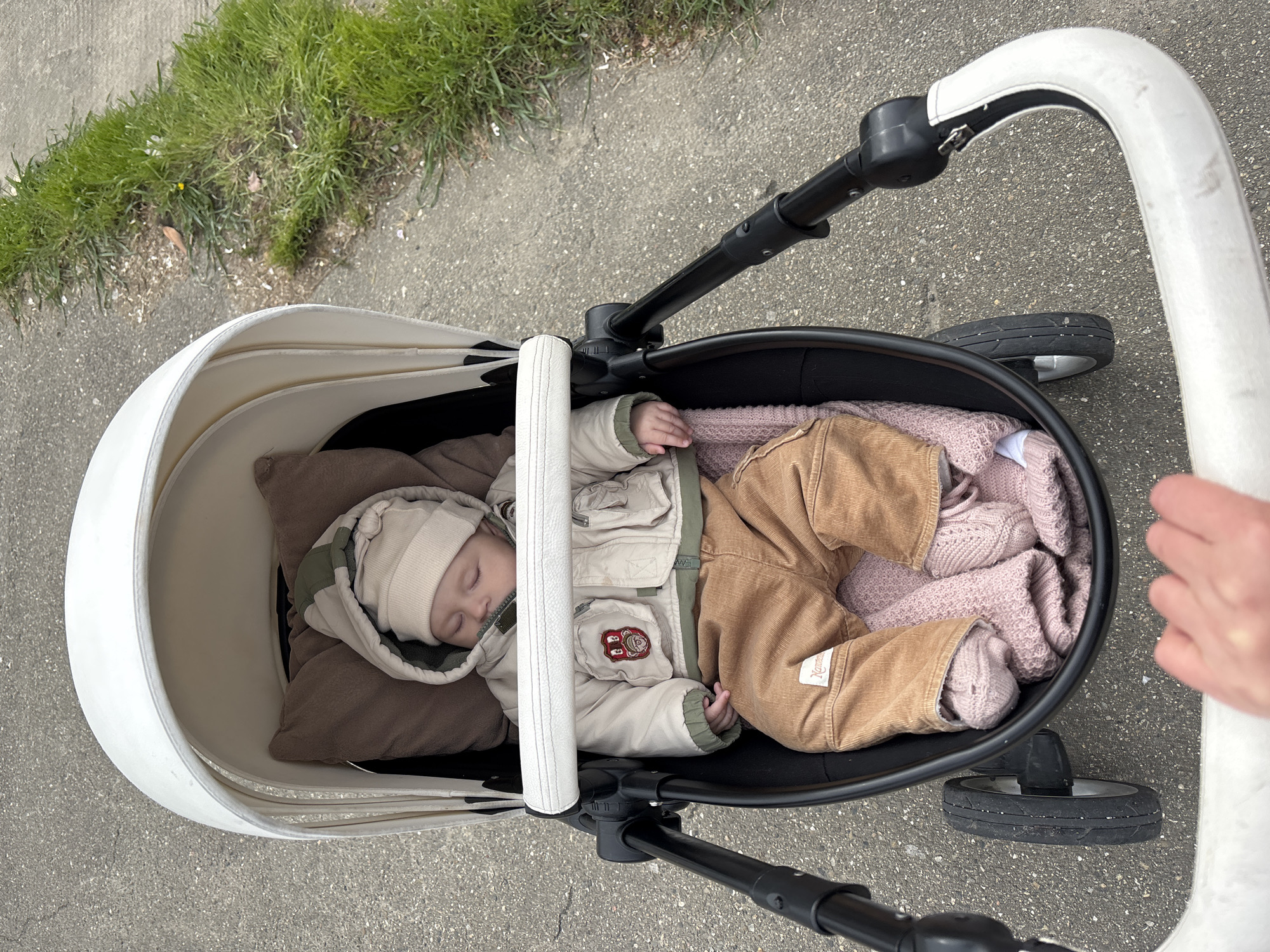 A baby is asleep in a stroller. The stroller is black and white, with a white handle. The baby is wearing a beige hat, a beige jacket with green accents, and brown corduroy pants. The baby is lying on a brown pillow and covered with a pink blanket. The stroller is on a gray paved surface, next to a patch of green grass. An arm and hand are visible holding the stroller handle. The location is a city street, but specific details to confirm Sochi, Russia, are not visible in the image.