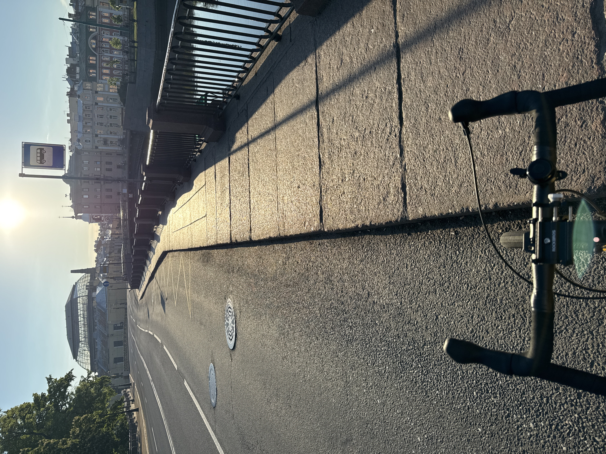 The image, taken at what appears to be either early morning or late afternoon, shows a scene along a paved embankment next to a body of water. In the foreground, the handlebars and a mount with the text "ROCKBROS" are visible from a bicycle, suggesting a first-person perspective from someone on a bike. Long shadows stretch across the textured stone wall of the embankment and the adjacent asphalt road, which features white lane markings and two circular manhole covers.
To the left of the road, an ornate black metal railing runs along the edge of the embankment, overlooking the water. Across the water, a row of classical European-style buildings with multiple stories and decorative facades lines the distant bank. A prominent blue-and-white bus stop sign, featuring a white bus icon, is visible on a pole, brightly lit by the low sun, which creates a lens flare in the lower left of the frame. Further along the opposite bank, a large structure with a distinctive glass and metal roof, possibly a historic market or exhibition hall, and a tall chimney or tower, stands out amidst other buildings and trees. The sky is clear, and the scene appears quiet and devoid of people or traffic, indicating a calm atmosphere. The strong sunlight and long shadows are notable details of the time of day.
