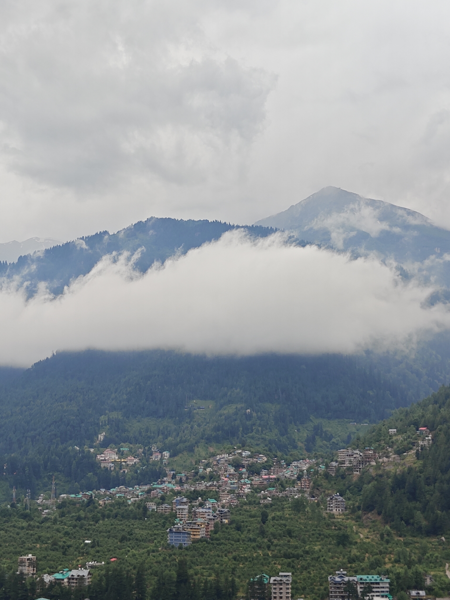 A medium-shot, high-angle, eye-level view captures the mountainous landscape of Vashisht, India, under a cloudy sky. Lush green trees cover the lower slopes of the mountains, giving way to a dense village settlement dotted with multi-story buildings. A thick band of white clouds partially obscures the middle section of the mountains, creating a sense of depth and mystique. The peak of a large mountain is visible in the background, its summit shrouded in mist. The village exhibits a mix of architecture, with some buildings featuring traditional sloped roofs and others displaying modern designs. The overall scene is one of natural beauty intertwined with human habitation.
