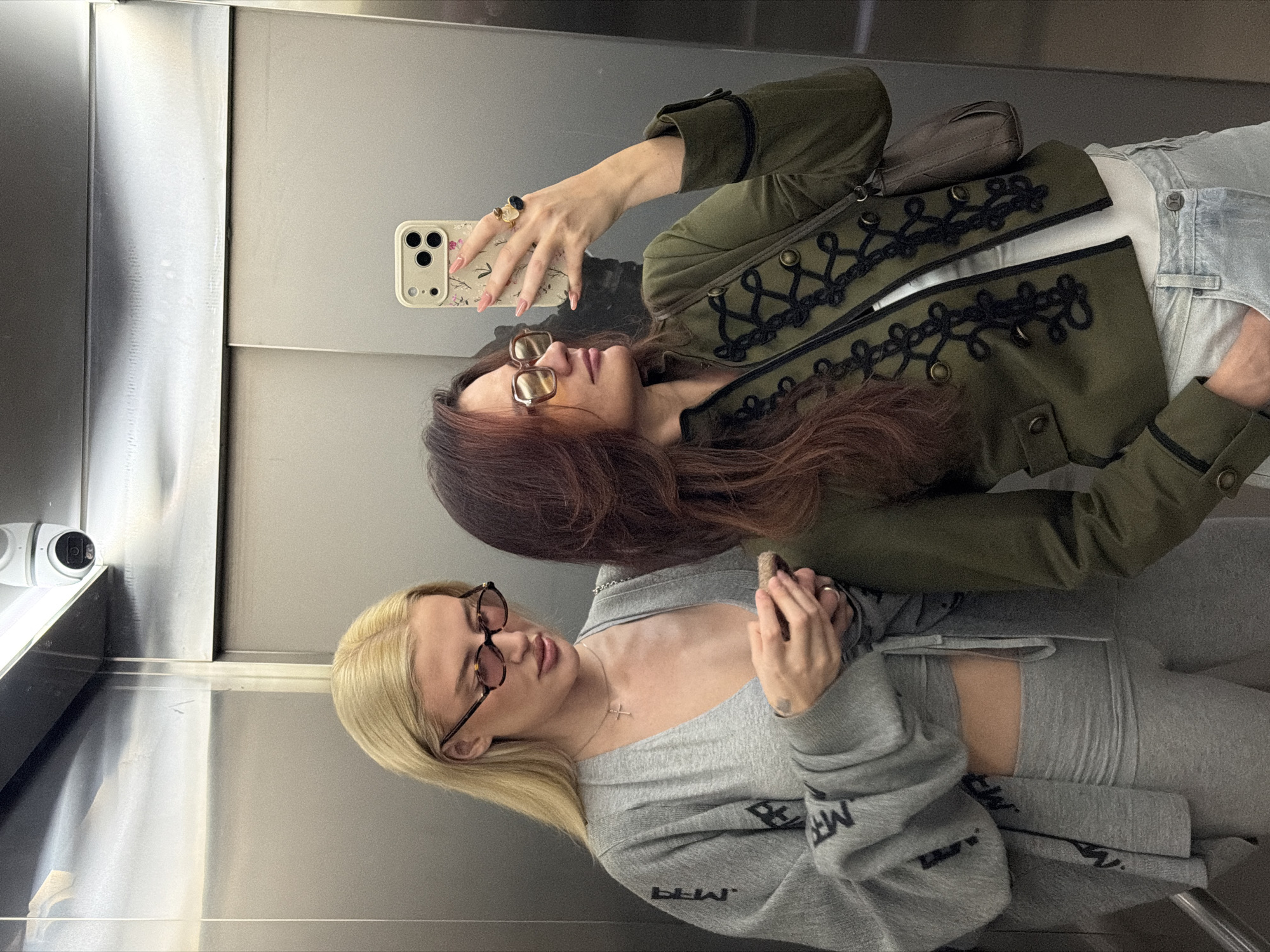 Two women are positioned in what appears to be the reflection of an elevator. The woman on the top, with dark hair and eyeglasses, is holding a smartphone taking a photo. She is wearing an olive green jacket with decorative black trim, a white top, and light blue jeans. She is wearing rings on multiple fingers. The woman below has blonde hair and eyeglasses. She is wearing a grey sweatshirt and matching pants. A security camera is visible in the upper left corner of the elevator. The elevator itself is metal. The location is not discernible from the image.