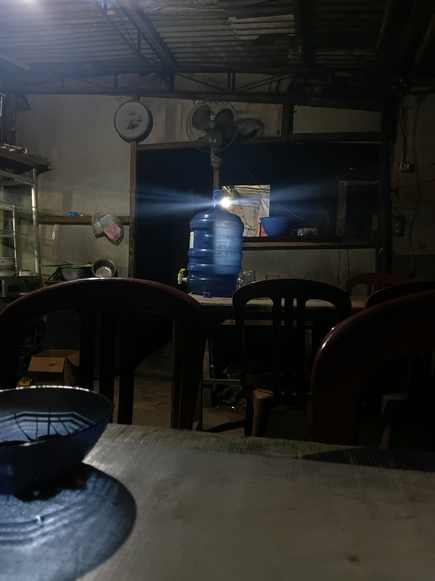 The image depicts an indoor scene, likely a food stall or small restaurant, dimly lit by artificial light. In the background, a large blue water dispenser is prominently placed on a table. To its left, a fan is visible, and further left, a wall-mounted scale is present. The foreground is dominated by a table and chairs, suggesting a seating area. The overall impression is one of a humble, functional establishment. The image was captured in Bantarbarang, Indonesia.
