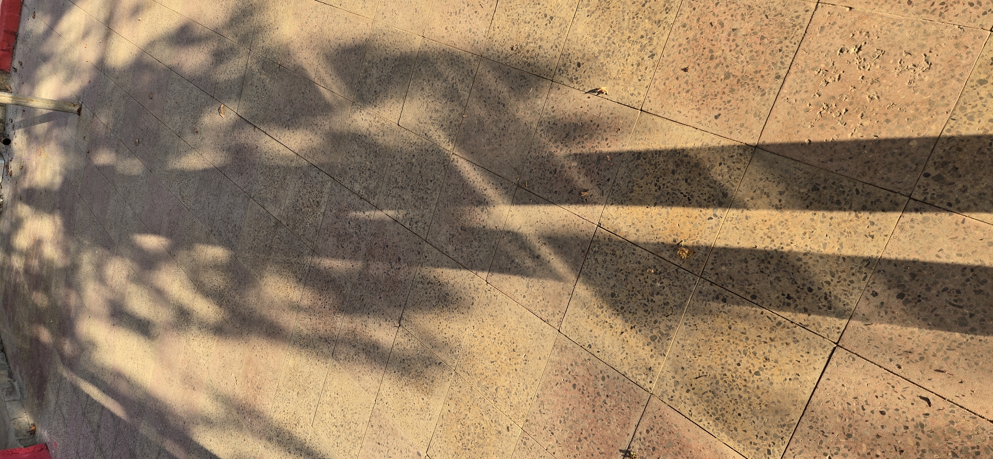 The image depicts a paved walkway with square tiles, casting long shadows from unseen objects, possibly trees or structures. The shadows are cast by sunlight filtering through foliage, creating an dappled effect on the ground. The tiles appear weathered and have a speckled pattern of light brown and gray. In the upper right portion of the image, a slender, light-colored pole or post is visible, casting a distinct shadow. The scene suggests an outdoor urban environment, possibly a park or a sidewalk in a city. There are no people visible in the image. The lighting indicates it is either early morning or late afternoon. The location is specified as Tel, Azerbaijan.