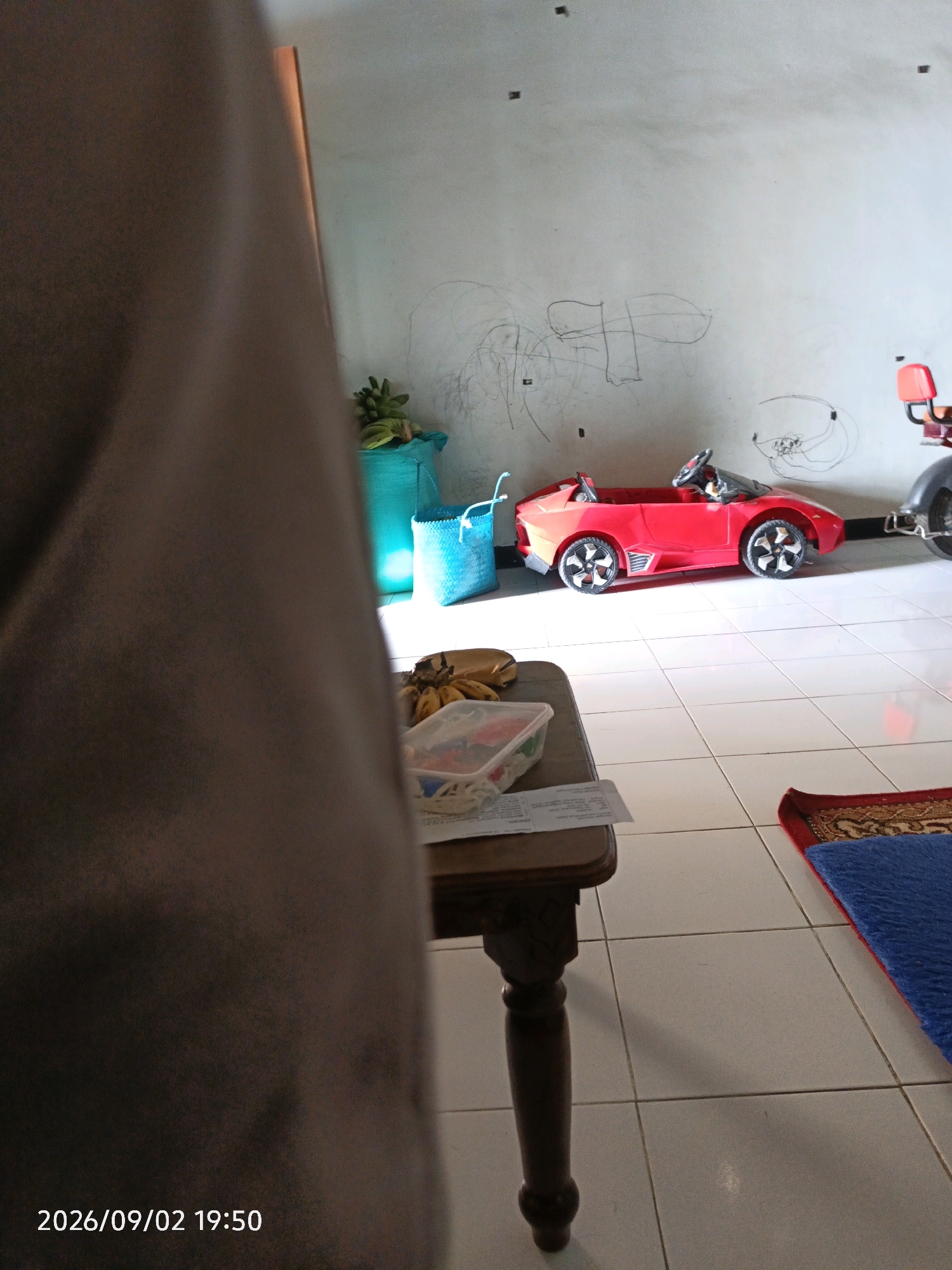 The image depicts an indoor scene with a tiled floor and a textured wall, likely in a residential setting in Tebuwung, Indonesia. In the foreground, a portion of a dark brown fabric drapes over the left side of the frame. A dark wooden table with carved legs stands in the mid-ground, displaying a plastic container filled with various items and a bunch of bananas. To the right of the table, a red toy car with black and silver wheels is positioned on the floor. Behind the toy car, a blue woven basket and a teal-colored bag are visible. The wall in the background is marked with chalk drawings, suggesting children's activity. Sunlight streams in from the right, casting shadows on the floor. A corner of a rug, featuring red, gold, and blue patterns, is visible in the lower right portion of the image. The timestamp "2026/09/02 19:50" is present in the lower left corner.