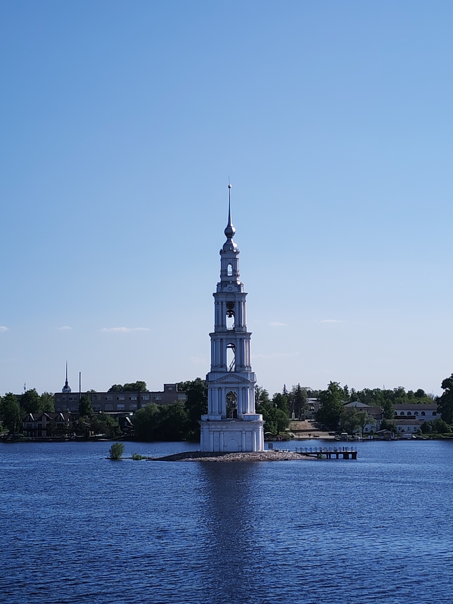 The image depicts the Kalyazin Bell Tower, a Neo-Renaissance Orthodox church tower located on a small island in the Uglich Reservoir, near Kalyazin, Russia. The tower, a prominent landmark, rises vertically from the center of the island, surrounded by blue water under a clear sky. The background shows a cityscape with trees and various buildings along the shoreline. A small dock extends into the water from the island near the tower.