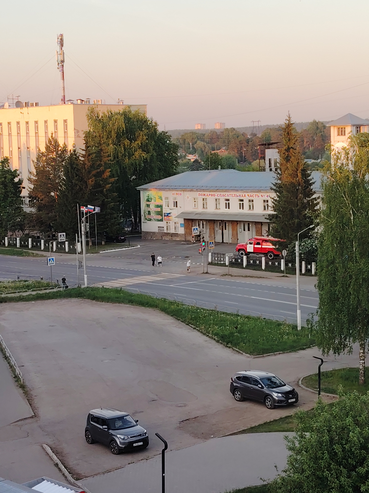 The image depicts a view from above, capturing a street scene in what is identified by the building signs as the city of Chaikovsky, Russia. The central focus is on a two-story building labeled "FIRE AND RESCUE STATION No. 47". A vintage red fire truck is parked in front of the building.
The street is visible, with a crosswalk where several people are crossing. There are two cars parked in a lower foreground area that appear to be a parking lot. The car closer to the viewer is a dark gray Kia Soul, while the other is a Honda CRV.
In the background, there are more buildings, including a tall structure with a communications tower, and a more distant tree line on the horizon. The lighting suggests it is either early morning or late afternoon, as the light is soft and casting long shadows. The sky is clear and tinted with the colors of sunrise or sunset.