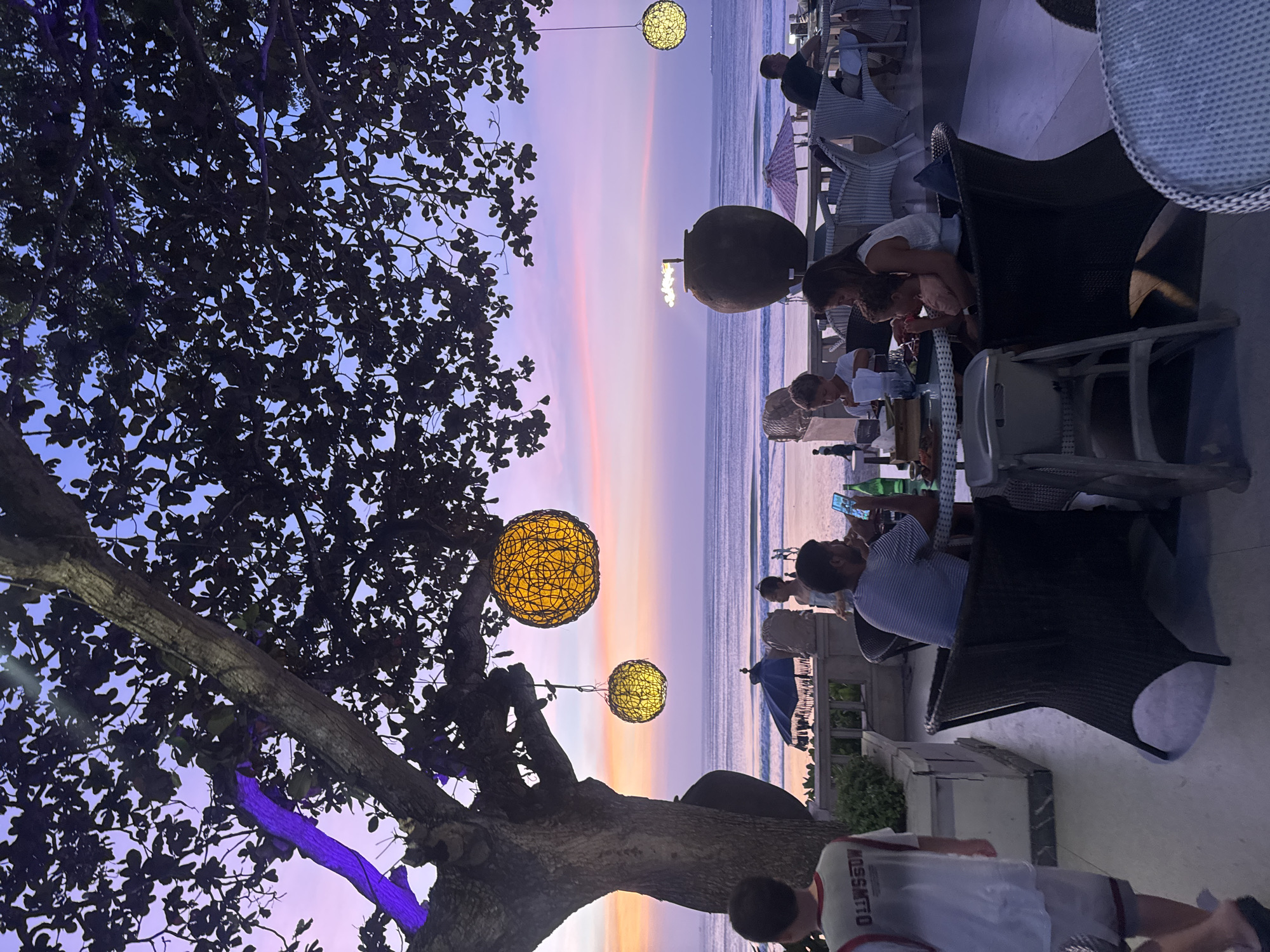 The image depicts an outdoor dining scene on a beach in Kuta Selatan, Indonesia, during a sunset. A large tree with dark foliage dominates the left side of the frame, with two illuminated spherical lanterns hanging from its branches. In the foreground, a group of people are seated at a table, seemingly enjoying a meal. A man in a striped shirt is looking at his phone, while a woman holds a child. To the left, a young person in a t-shirt walks away from the camera. The background shows the ocean with gentle waves and a colorful sky, transitioning from hues of purple to orange. Other diners and beachgoers are visible in the distance, along with umbrellas and more seating. The overall ambiance is tranquil and picturesque.