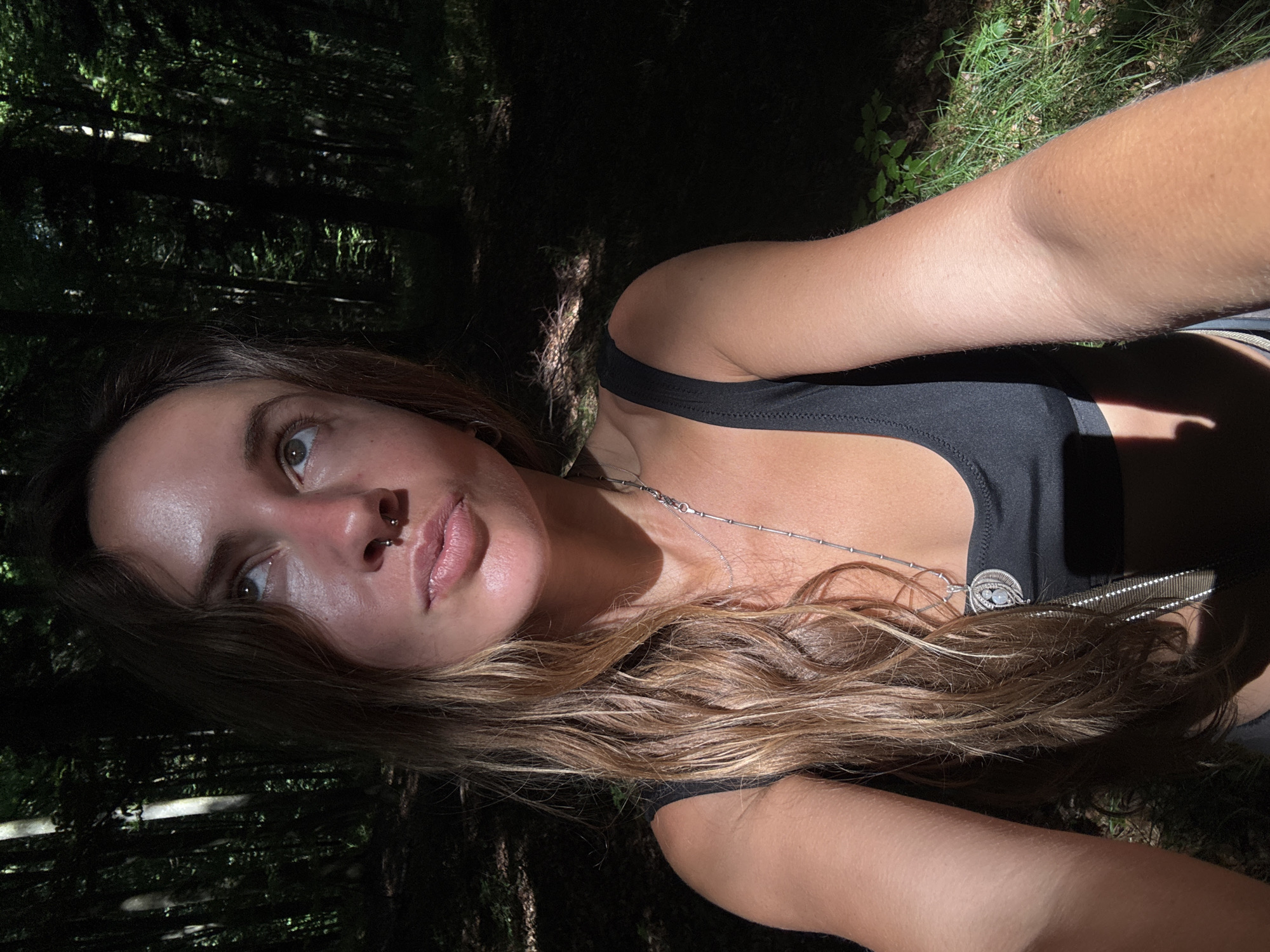 This is a self-portrait of a young woman taken outdoors. She is lying down in a wooded area, with dark trees and some grassy ground visible in the background. The lighting suggests it is daytime, possibly late afternoon, as there are strong shadows and dappled sunlight. The woman has long, wavy brown hair and is wearing a black tank top. She is looking up and to her left, with a neutral expression. A silver necklace with a circular pendant is visible around her neck. A nose ring is also visible. The image is oriented sideways.