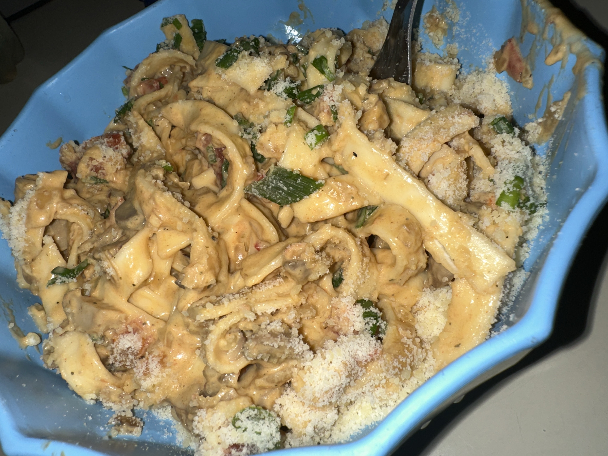 The image displays a close-up view of a pasta dish served in a light blue, fluted-edge bowl. The bowl is filled with a creamy, light brown sauce coating broad, flat pasta noodles that appear to be folded or rolled. Mixed into the pasta are chopped green ingredients, likely scallions or chives, along with small red pieces that could be diced tomatoes or red bell pepper, and possibly mushrooms. The entire dish is generously topped with a substantial amount of finely grated cheese, resembling Parmesan, which also spills onto the inner rim of the bowl. A silver fork is partially visible, inserted into the pasta on the right side of the bowl, suggesting the meal is either being eaten or has just been served. The lighting is dim, indicating an indoor setting, possibly an evening meal. No people or visible text are present. The context provided specifies the location as Buenos Aires, Argentina, though there are no visual cues within the image to confirm this.