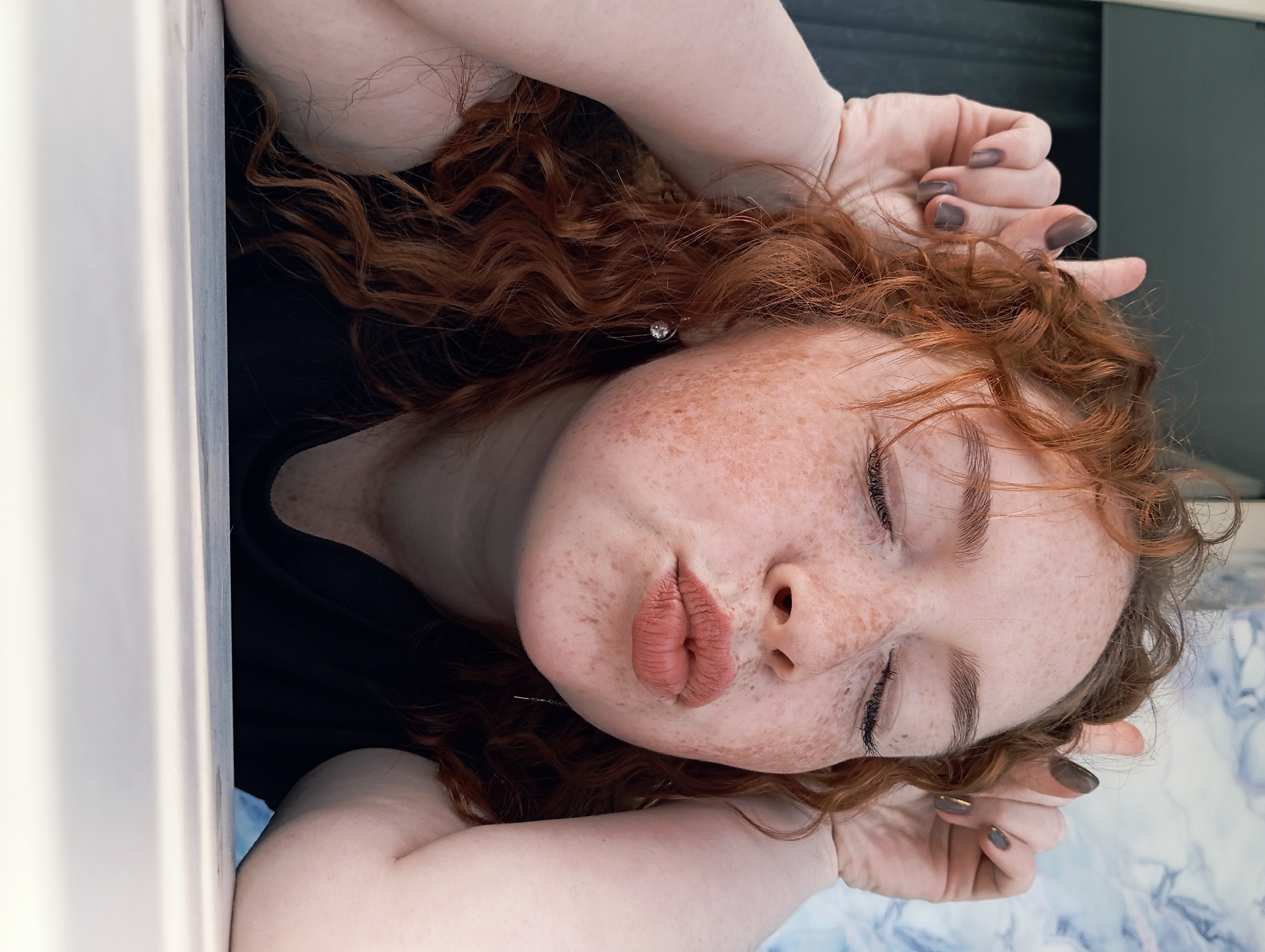 A young woman with red curly hair is lying down with her eyes closed. Her freckled face is tilted upward. Her hands are near her head, with her fingernails painted a dark color. She is wearing a black top. The background includes what appears to be a white surface and a mottled blue and white surface. It is difficult to determine the specific location of this image in Yurgamysh, Russia.