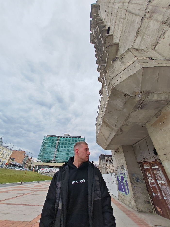 An adult male, approximately 20-30 years of age with short hair, stands on a paved surface in Shumen, Bulgaria. He is positioned in the lower-left quadrant, angled slightly right and gazing towards the right side of the frame. He wears a black hooded sweatshirt with "DYSTOPIA" printed in white on the front, underneath a dark overcoat.
To the immediate right of the male, a large, multi-tiered concrete structure, part of the Monument to 1300 Years of Bulgaria, rises prominently. This brutalist-style edifice exhibits weathered grey concrete, visible cracks, and exposed rebar, with sections of metal railing on an upper tier. Its lower section features graffiti in purple and green spray paint near a rusted, dark brown double-door entrance.
In the mid-ground, to the left of the monument, a multi-story building, identified as the Shumen Plaza (Hotel Shumen), is entirely enveloped in green scaffolding and netting, indicating ongoing construction or renovation. Further to the left, other urban buildings with classical architectural elements are partially visible. The ground beneath the male is composed of light-colored geometric pavers. The sky overhead is uniformly overcast with a grey cloud cover.