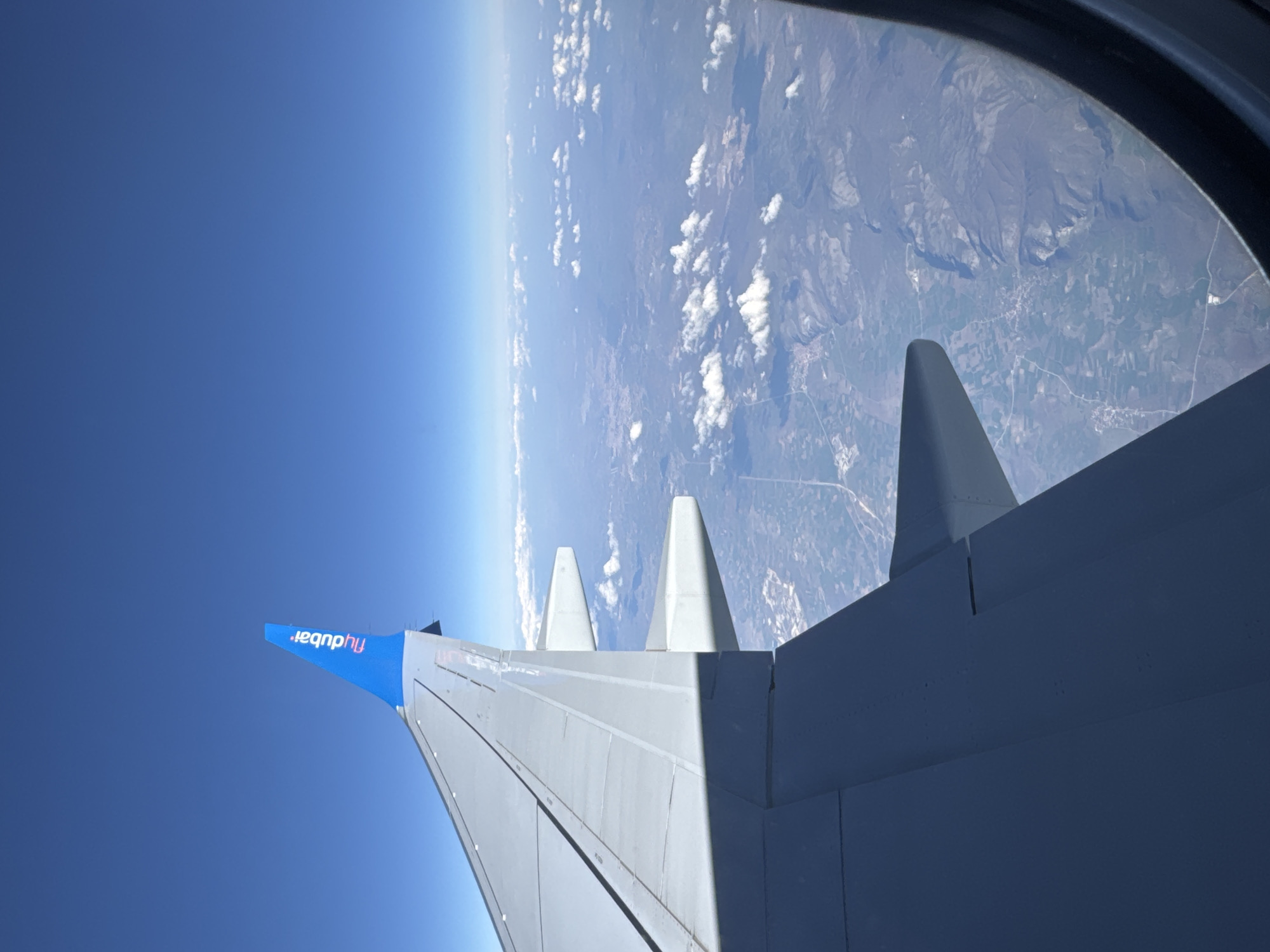 A view from an airplane window shows a wing with the "flydubai" logo against a blue sky. The Earth is visible in the distance, with clouds and land. It is unclear if Burgas, Bulgaria is in the image, given the limited visible information.