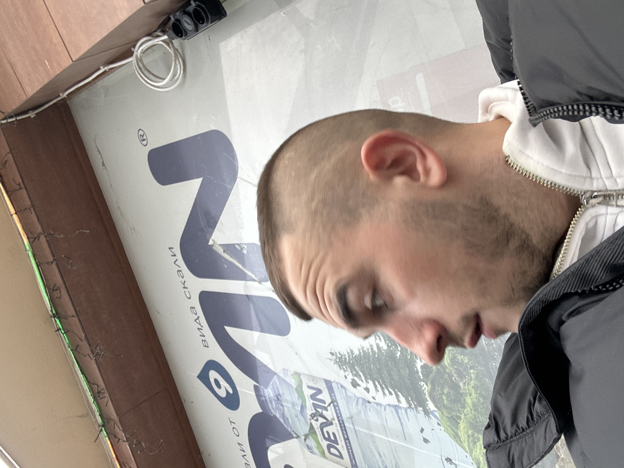 A low-angle, close-up shot captures a man standing in front of an advertisement. The man has short, buzzed hair, a beard, and is wearing a black jacket over a white hoodie. He is looking down, partially obscuring his face.
Behind him is a large advertisement in Bulgarian with the brand name "Devin" prominently displayed. The advertisement features a mountain landscape. The words "вида скали" ("view of the rocks") are visible on the sign.
The location is in Plovdiv, Bulgaria, as evidenced by the Bulgarian writing on the advertisement. The presence of electrical wiring and a brown wooden structure suggests an urban outdoor setting, possibly a storefront or bus stop.