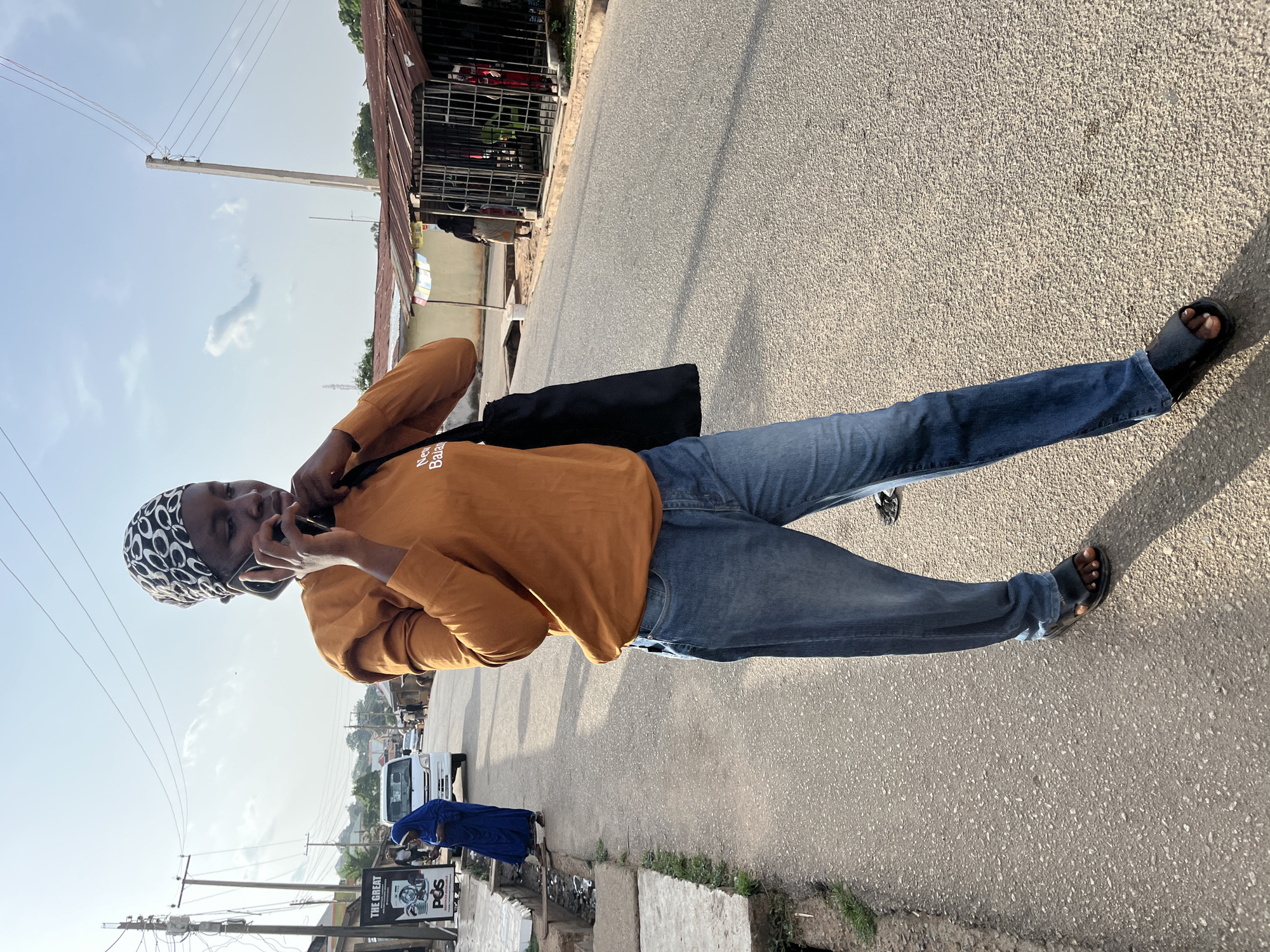 A person wearing jeans, an orange shirt, and a black-and-white head covering stands on an asphalt road, holding a phone to their ear. They also have a black shoulder bag. The setting is a street in Jos, Nigeria. A woman in a blue dress is visible near a sign that reads "The Great POS" and shows an ATM card. Buildings, electrical poles, and vehicles are present along the street.