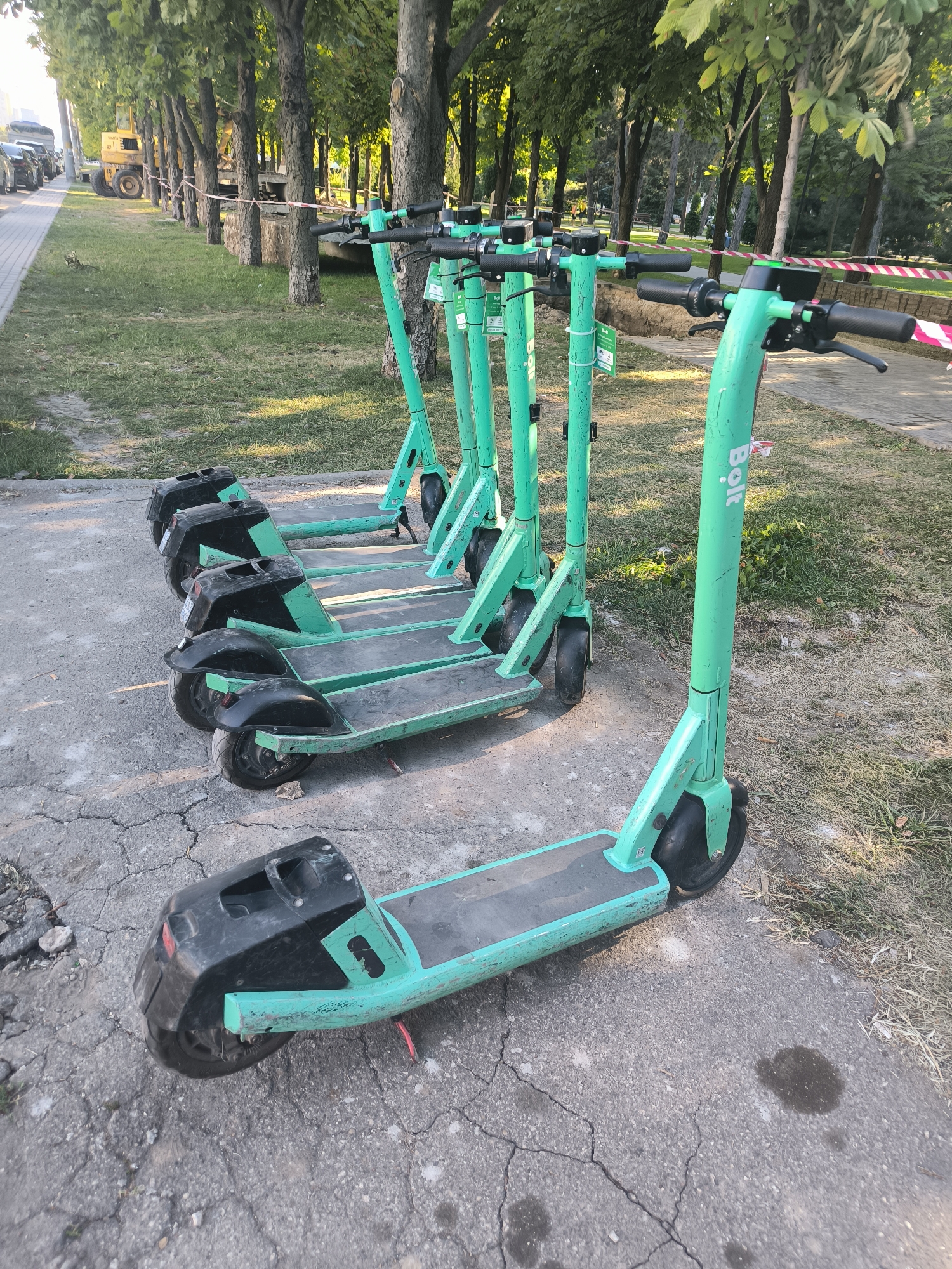 The image depicts a row of several green electric scooters parked on a paved surface next to a grassy area with trees. The scooters are facing towards the right side of the frame, with their handlebars visible. Some of the scooters have "Bolt" branding on their frames. In the background, there are more trees and a red and white striped tape barrier. To the left of the scooters, there is a sidewalk with some construction equipment visible, including a yellow excavator. The scene appears to be in an urban environment, possibly a park or a street in Chișinău, Moldova, given the context. The lighting suggests it is daytime, possibly late afternoon or early morning due to the long shadows. There are no people visible in the image.