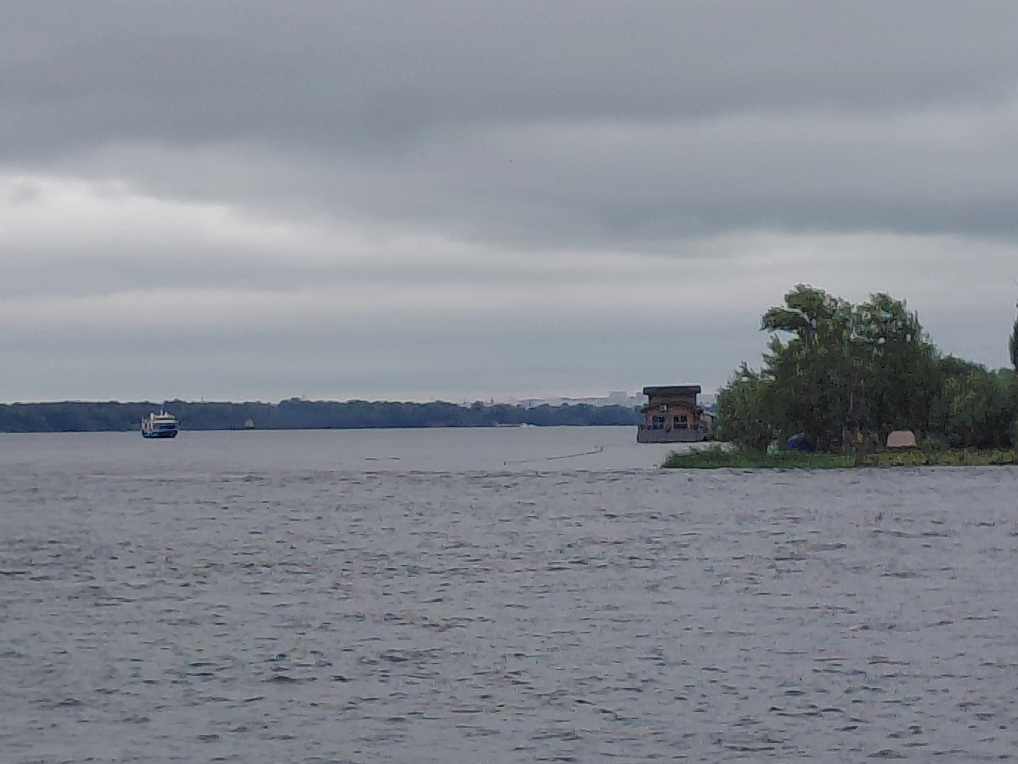 An overcast, wide shot captures a body of water with a distant tree line under a cloudy sky. On the left side of the water, a blue and white ferry is visible. On the right side of the water, a multi-story houseboat with a brown roof sits on a patch of green land, with tents and trees nearby. The image appears to be taken from a boat or the shore, with the water showing ripples. The location is the Volga River in Novokuybyshevsk, Russia.