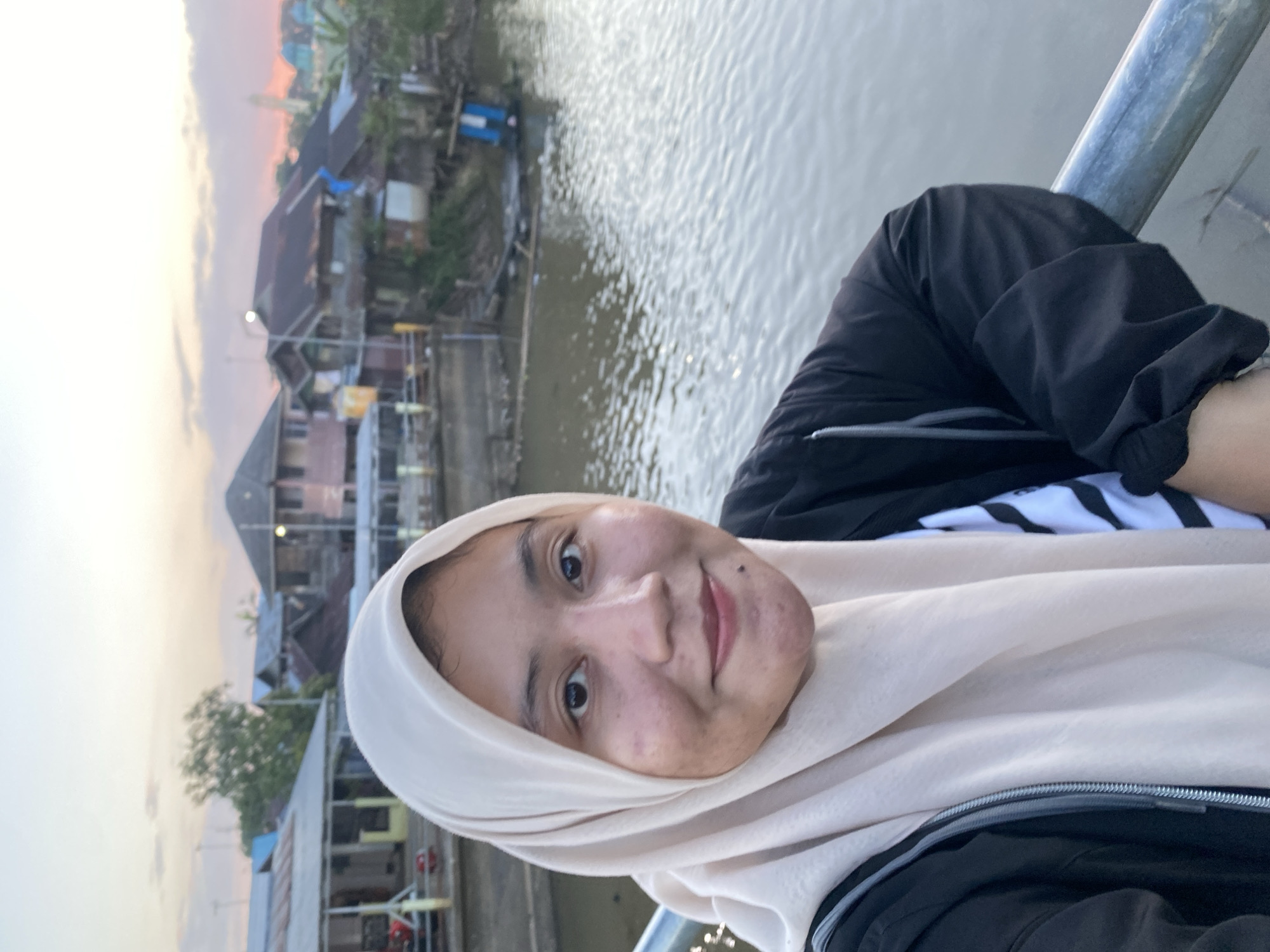 A young woman wearing a pale beige hijab and a black zip-up jacket over a striped shirt takes a selfie on a bridge. She is smiling and looking at the camera. In the background, there is a river and a village with wooden houses on stilts. The sky is a soft pink and orange, indicating it is either sunrise or sunset. The location is Tunggul Irang Ulu, Indonesia.