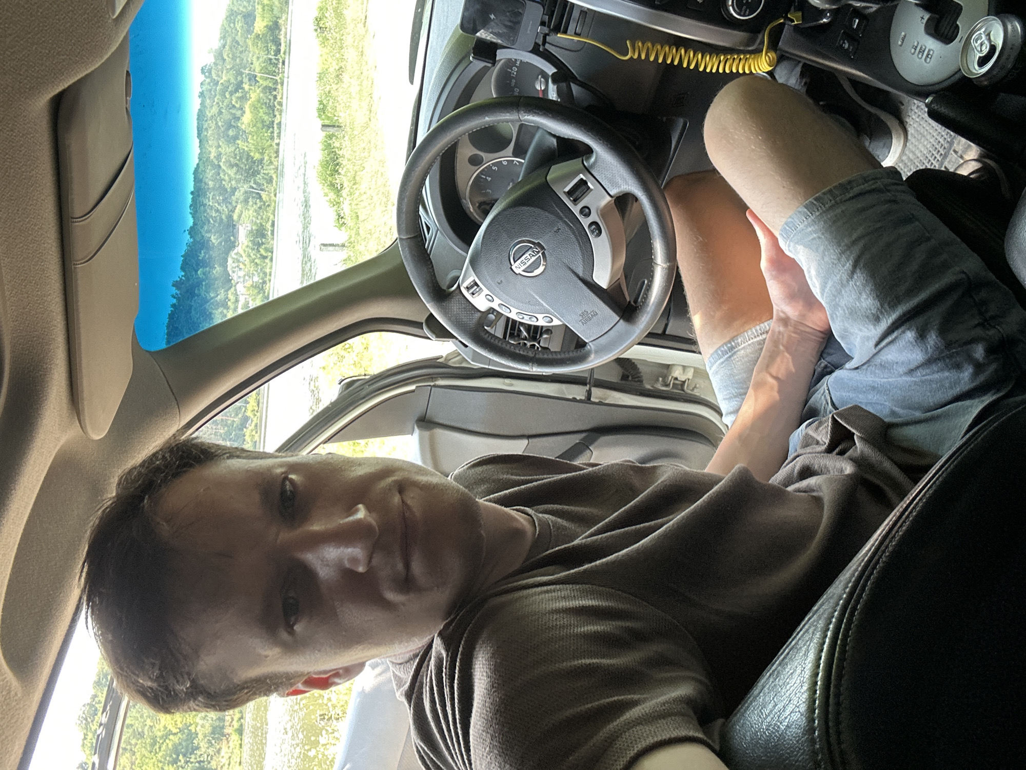 A man is sitting in the driver's seat of a car, looking towards the camera. The car is parked outdoors, with a body of water and lush green hills visible through the windshield. The man is wearing a brown t-shirt and denim shorts. The interior of the car shows a steering wheel with a Nissan logo, dashboard, and gear shift. A bright yellow coiled cable is attached to the dashboard. The scene suggests a scenic location, possibly a park or a roadside with a view. The location is identified as Makiv, Ukraine.