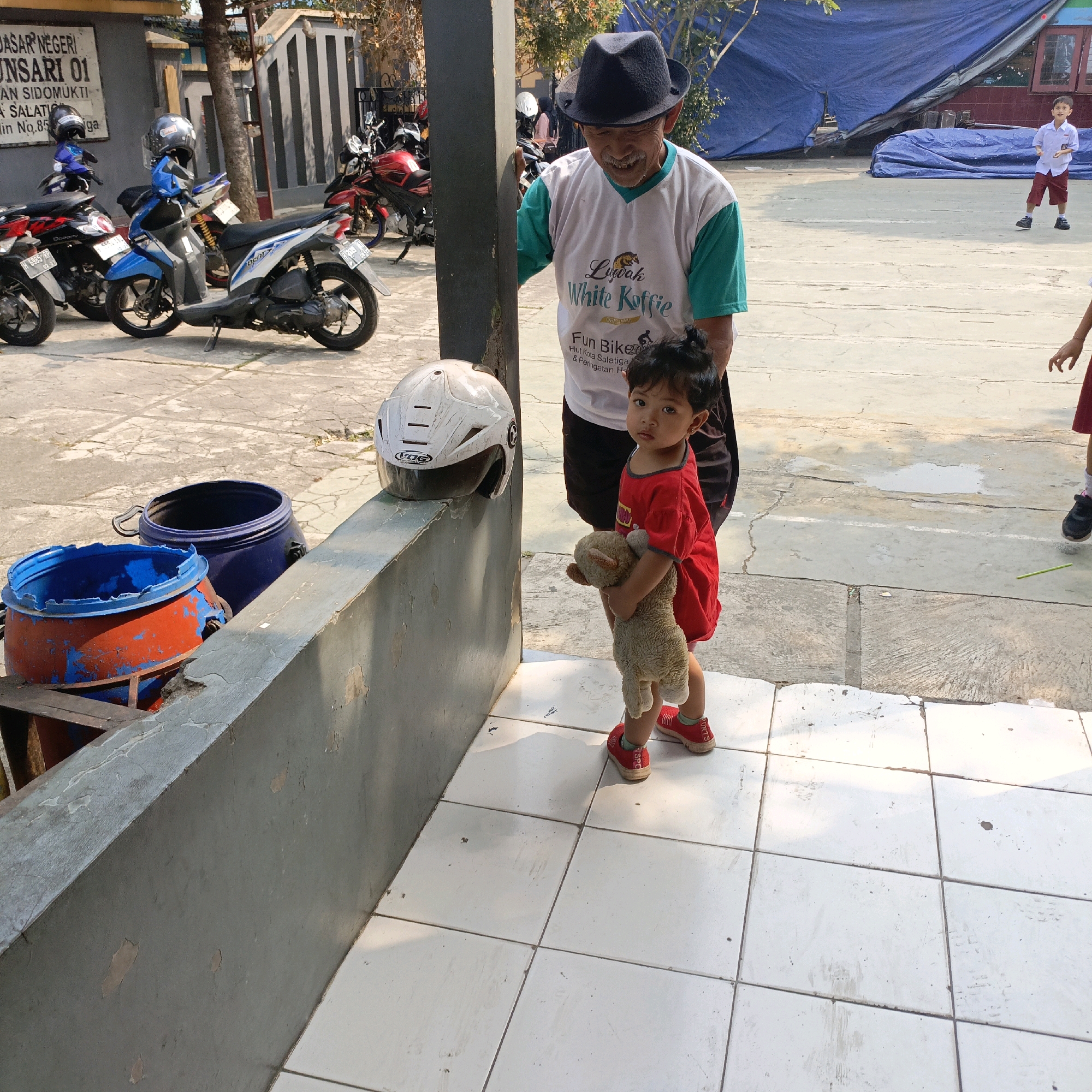 In front of a building with a sign that reads "DASAR NEGERI SUNSARI 01" and "Jl. Sidomukti", a man wearing a black fedora and a white t-shirt with green sleeves stands looking down at a young child. The man has a mustache and is smiling. The child, wearing a red shirt and red shoes, is holding a stuffed animal. A white helmet rests on a low wall in the foreground. Several motorcycles are parked in the background. A blue tarp covers part of the building. The scene appears to be outdoors on a paved area.